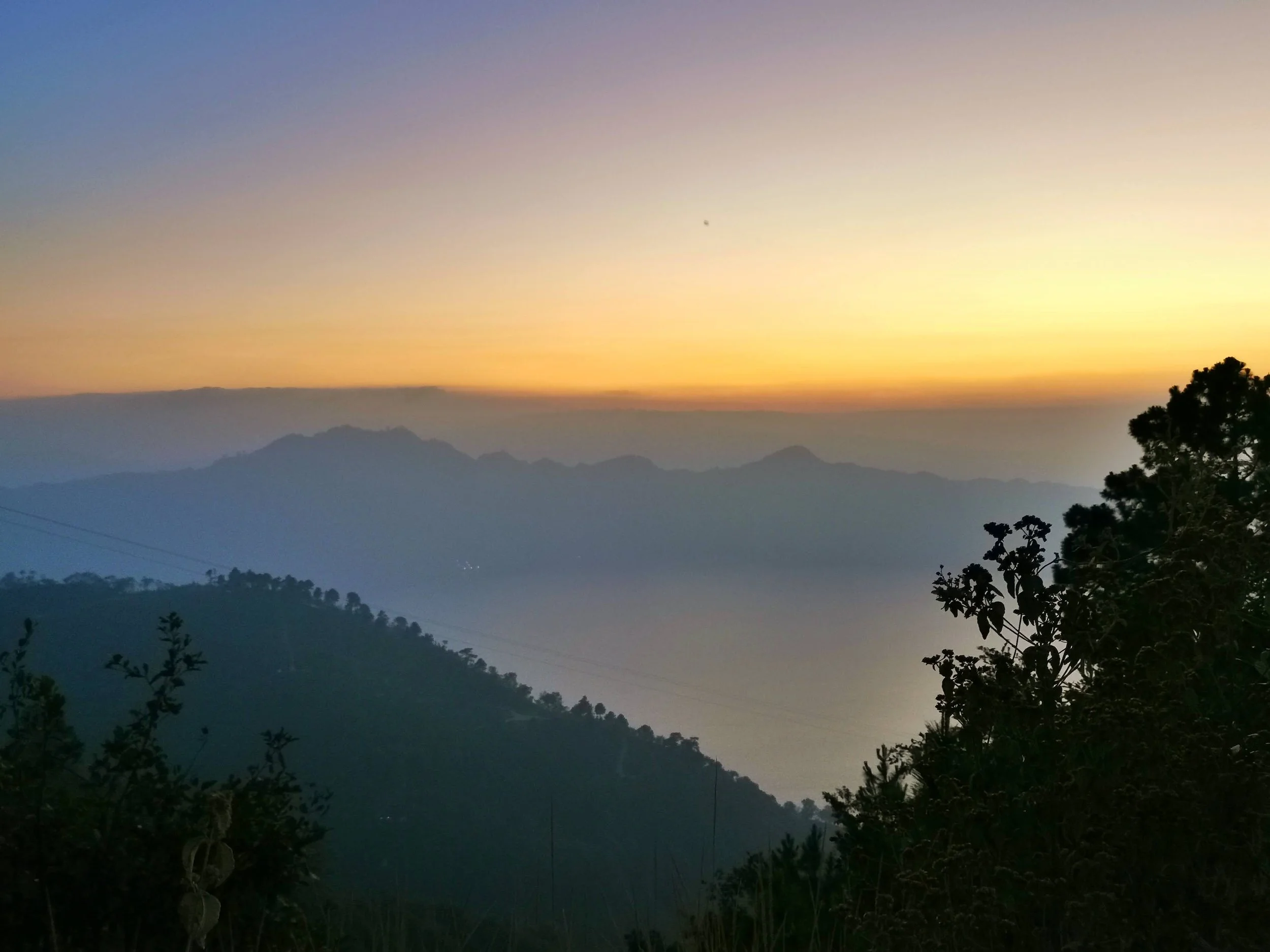 Sunset over mountain range with silhouettes of trees and faint moon in the sky.