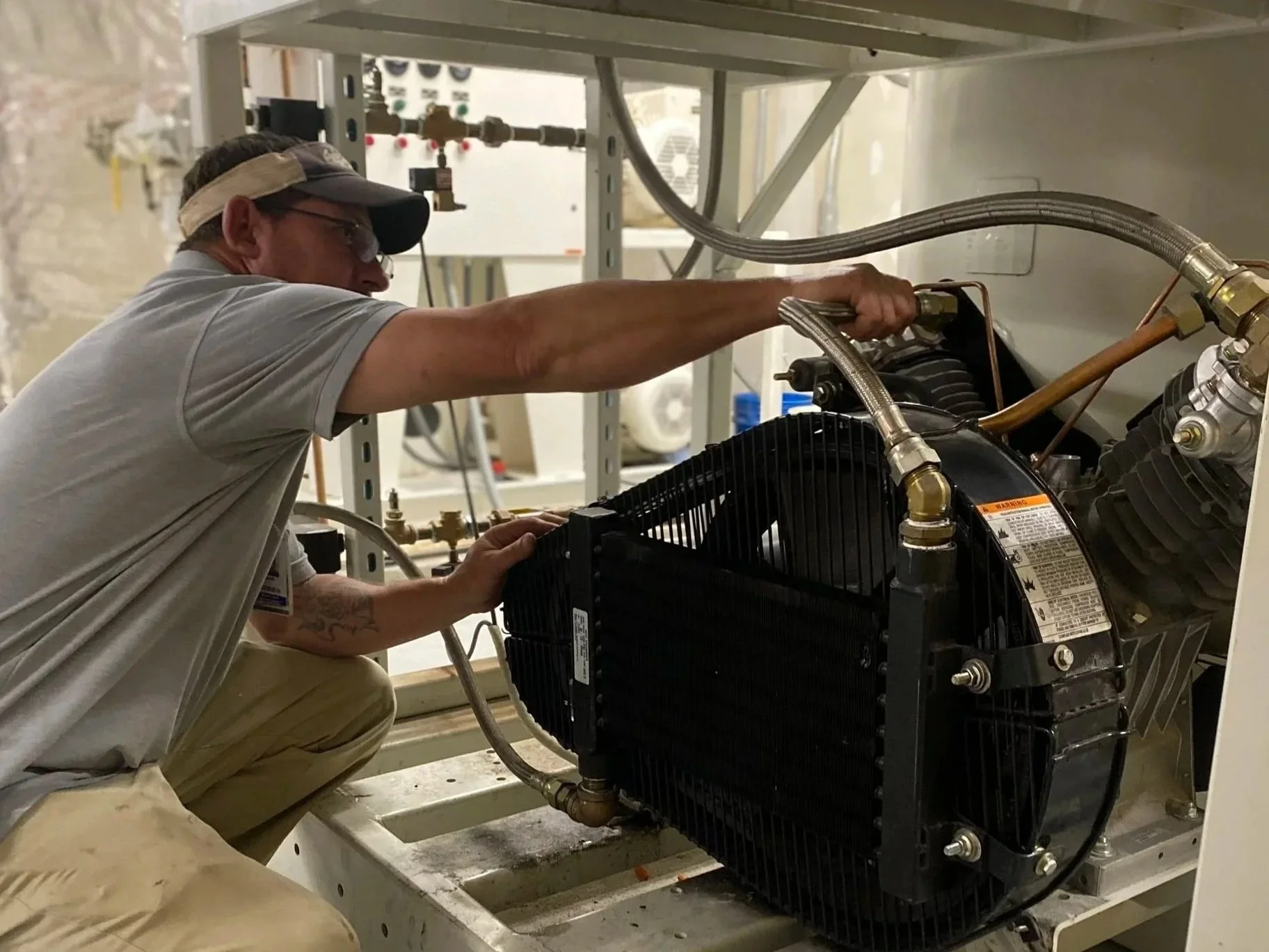 A technician working on an industrial piece of equipment inside a facility, wearing glasses, a beige cap, and casual clothes, adjusting or inspecting components with various pipes, wires, and a black cooling unit visible.