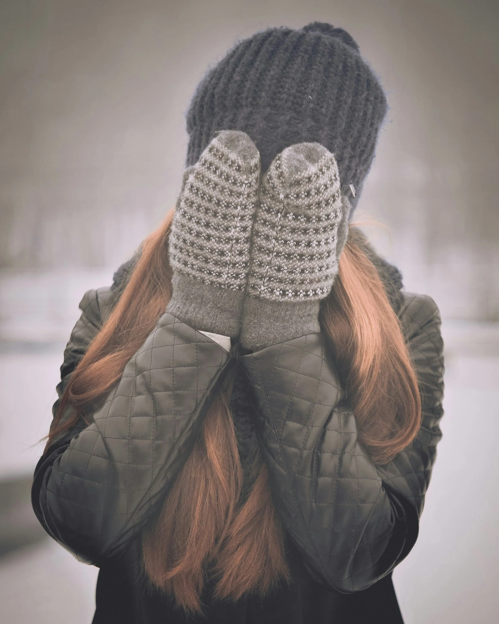 Person wearing a black knit beanie, gray mittens with snow, black quilted jacket, and with long brown hair, standing outdoors in winter, covering their face with gloved hands.