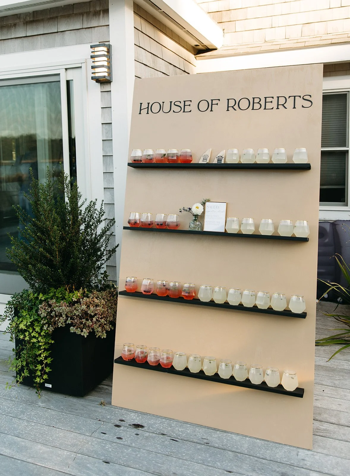 A large beige display board with the text 'HOUSE OF ROBERTS' at the top, featuring three black shelves holding glasses filled with various beverages in front of a house with an outdoor patio area, potted plants, and windows.