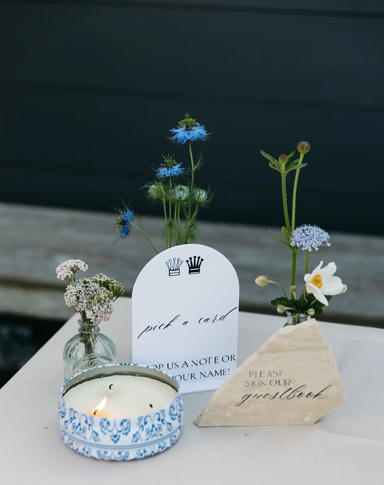 Table with small vases of wildflowers, a lit candle in a blue and white dish, and signs reading "pick a card" and "please sign our guestbook".