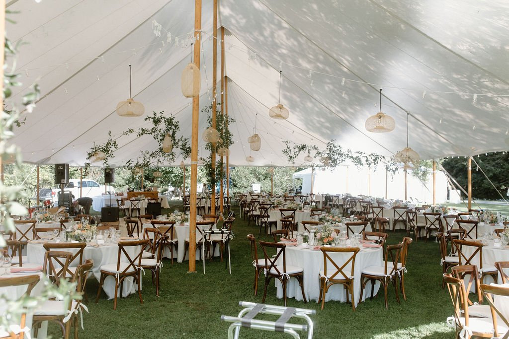 Wedding reception setup under a large white tent with round tables, wooden chairs, floral centerpieces, and hanging decorations.