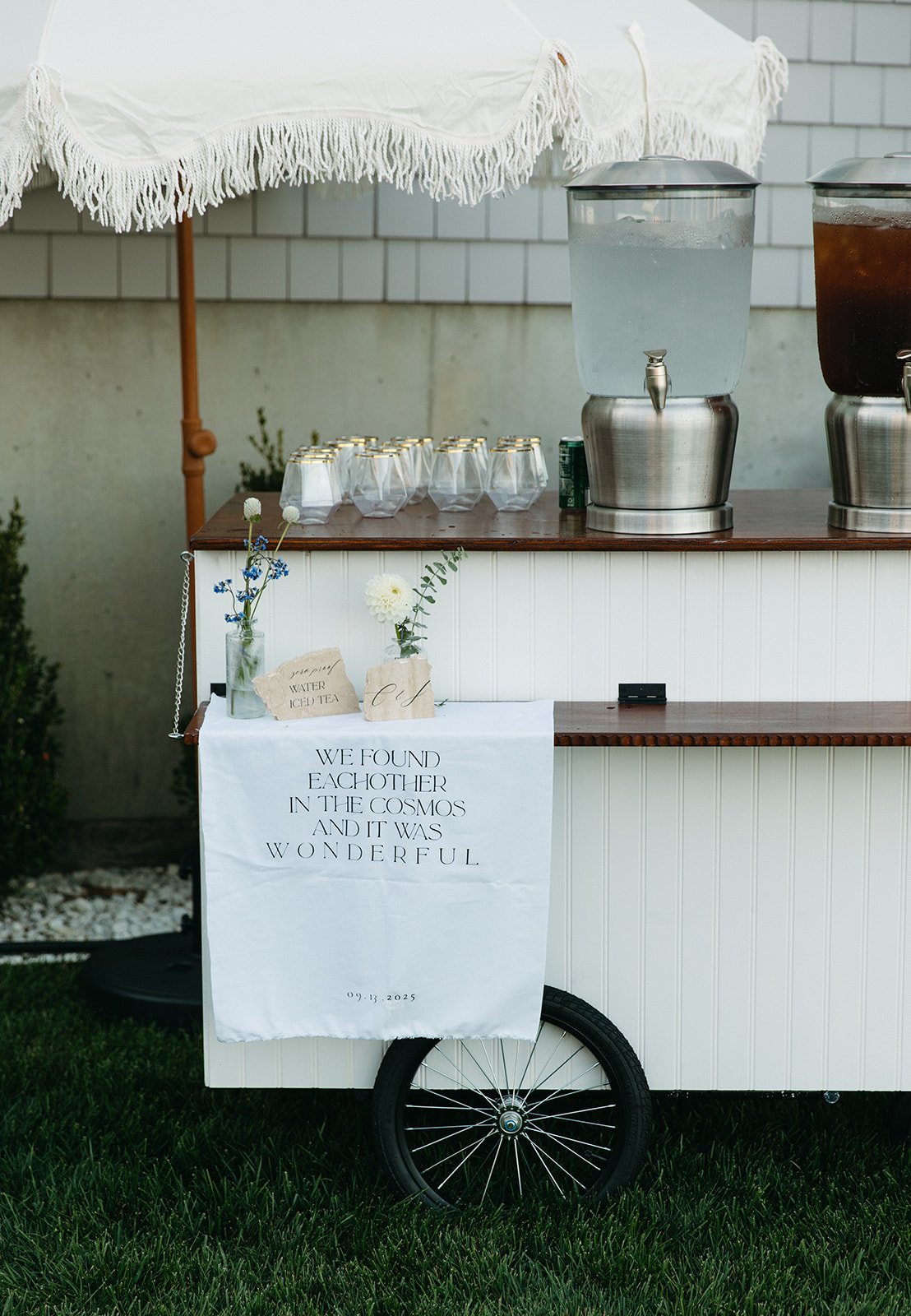 A beverage cart with two large beverage dispensers, glasses, and floral arrangements, set outdoors on grass near a white-paneled wall. A white cloth with a quote is draped over the front wheel of the cart.