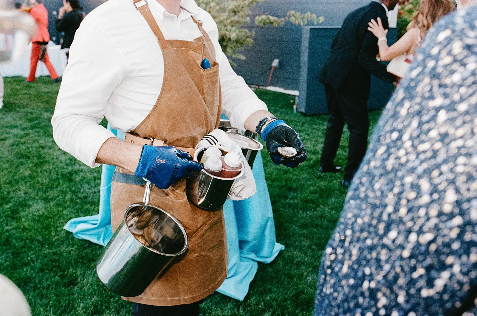 Close-up of a person wearing a brown apron and gloves, serving food or drinks at an outdoor event with other people dancing and socializing in the background.