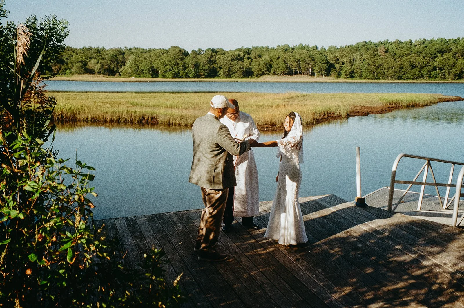 A couple getting married outdoors on a wooden dock by a lake, with a officiant conducting the ceremony, surrounded by nature and tall grass.