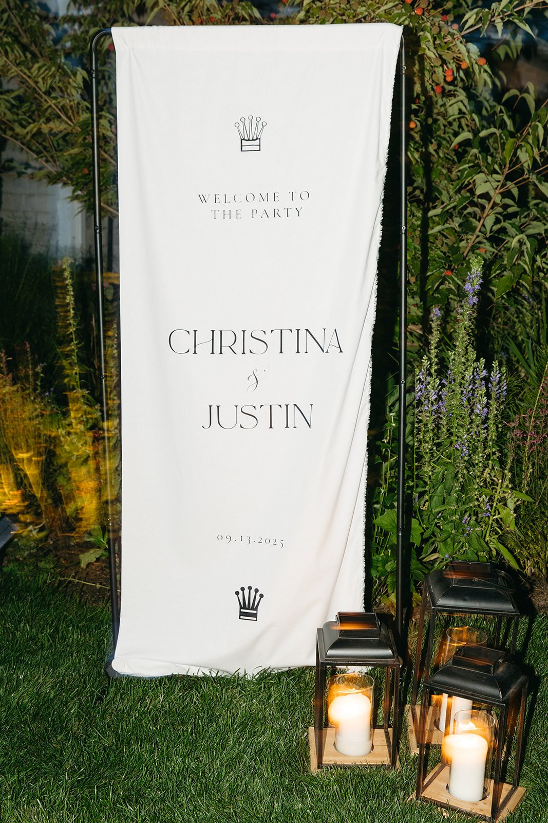 A wedding welcome sign with the names Christina and Justin and the date 09.13.2025, decorated with candles in lanterns and surrounded by greenery and flowers.