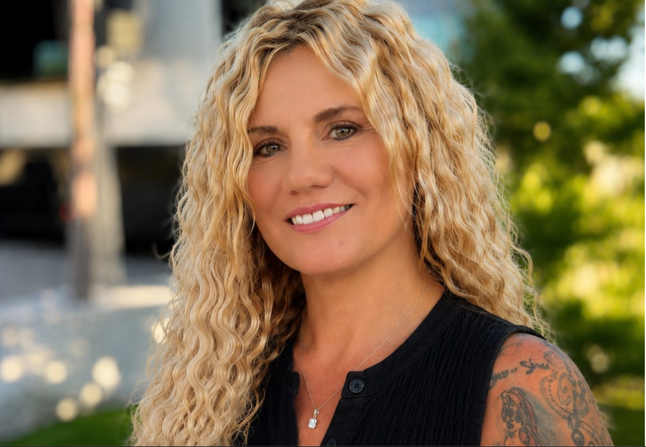 Close-up of a woman with long, curly blonde hair, smiling, outdoors with greenery in the background.
