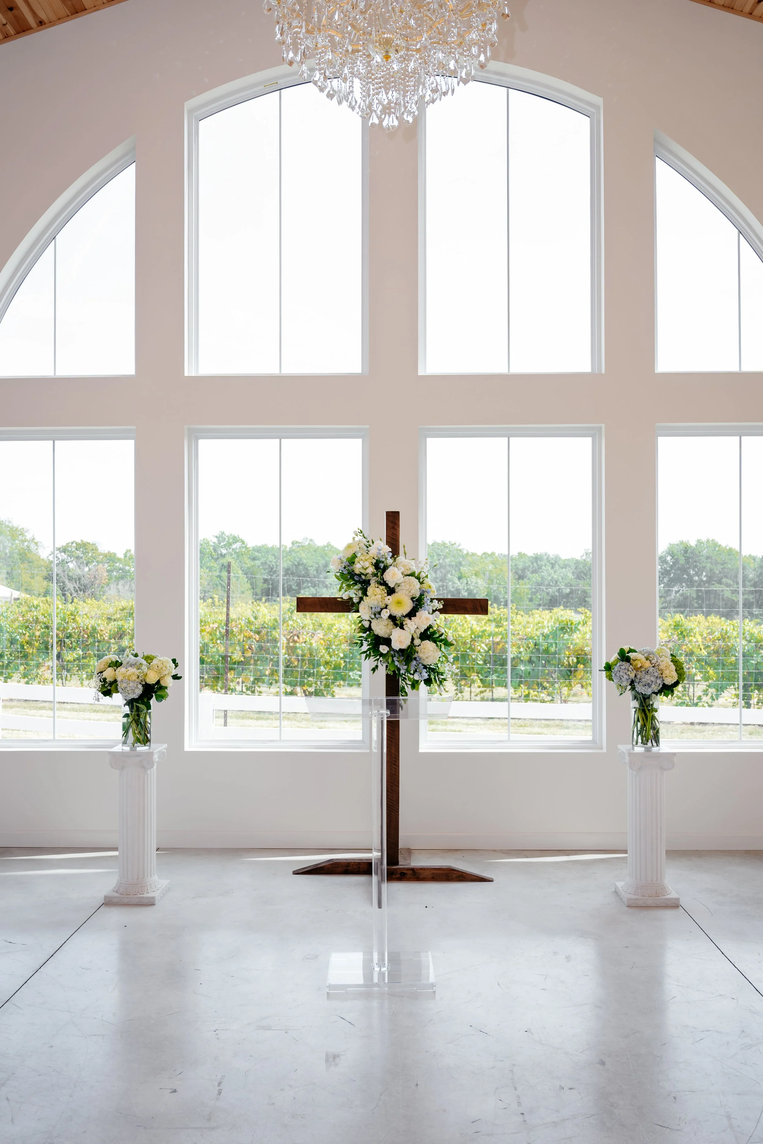 Interior of a wedding chapel with large windows, a wooden cross decorated with flowers, and two floral arrangements on white pedestal stands.