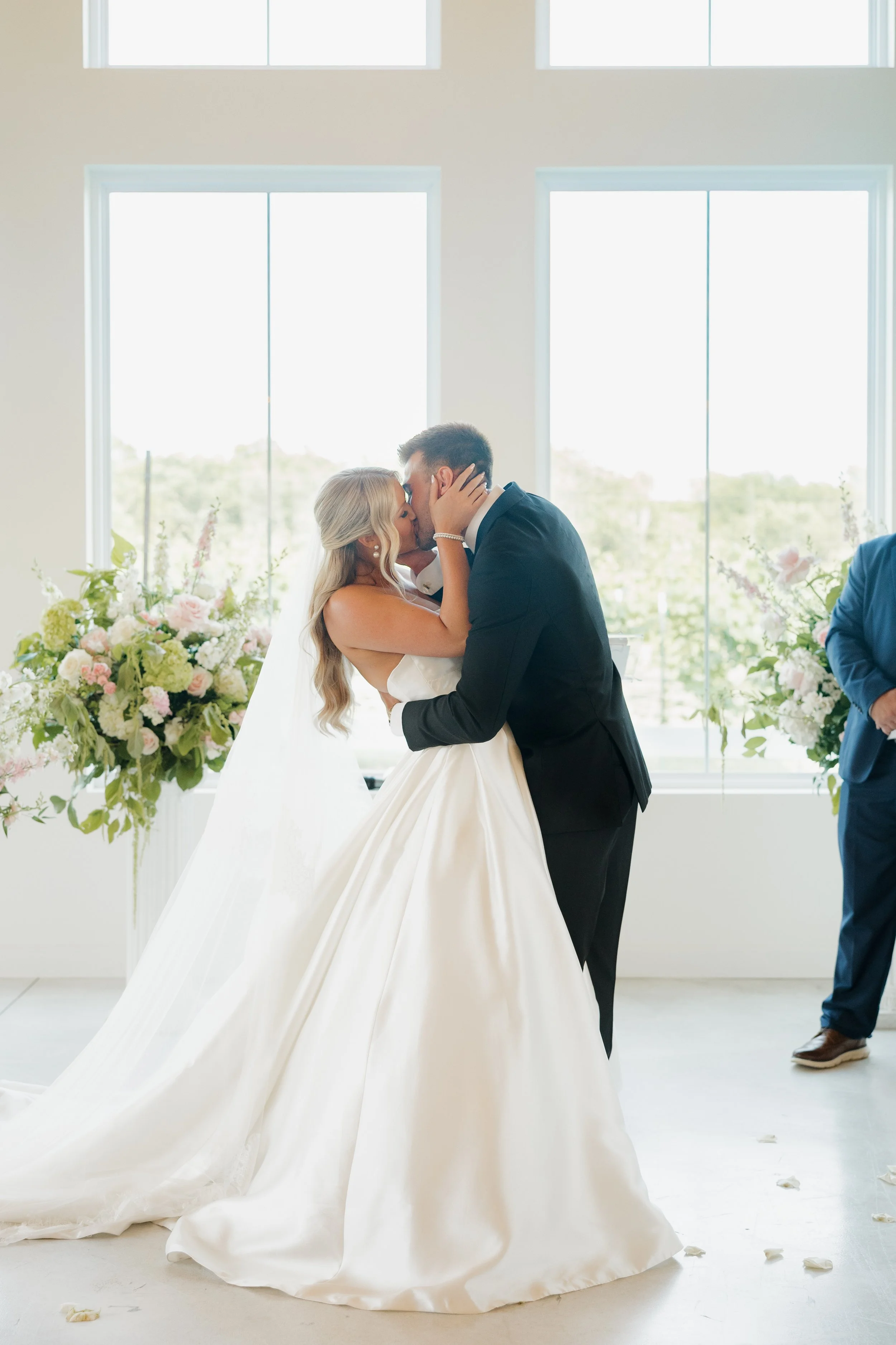 A bride and groom kissing during their wedding ceremony, with floral arrangements and large windows in the background.