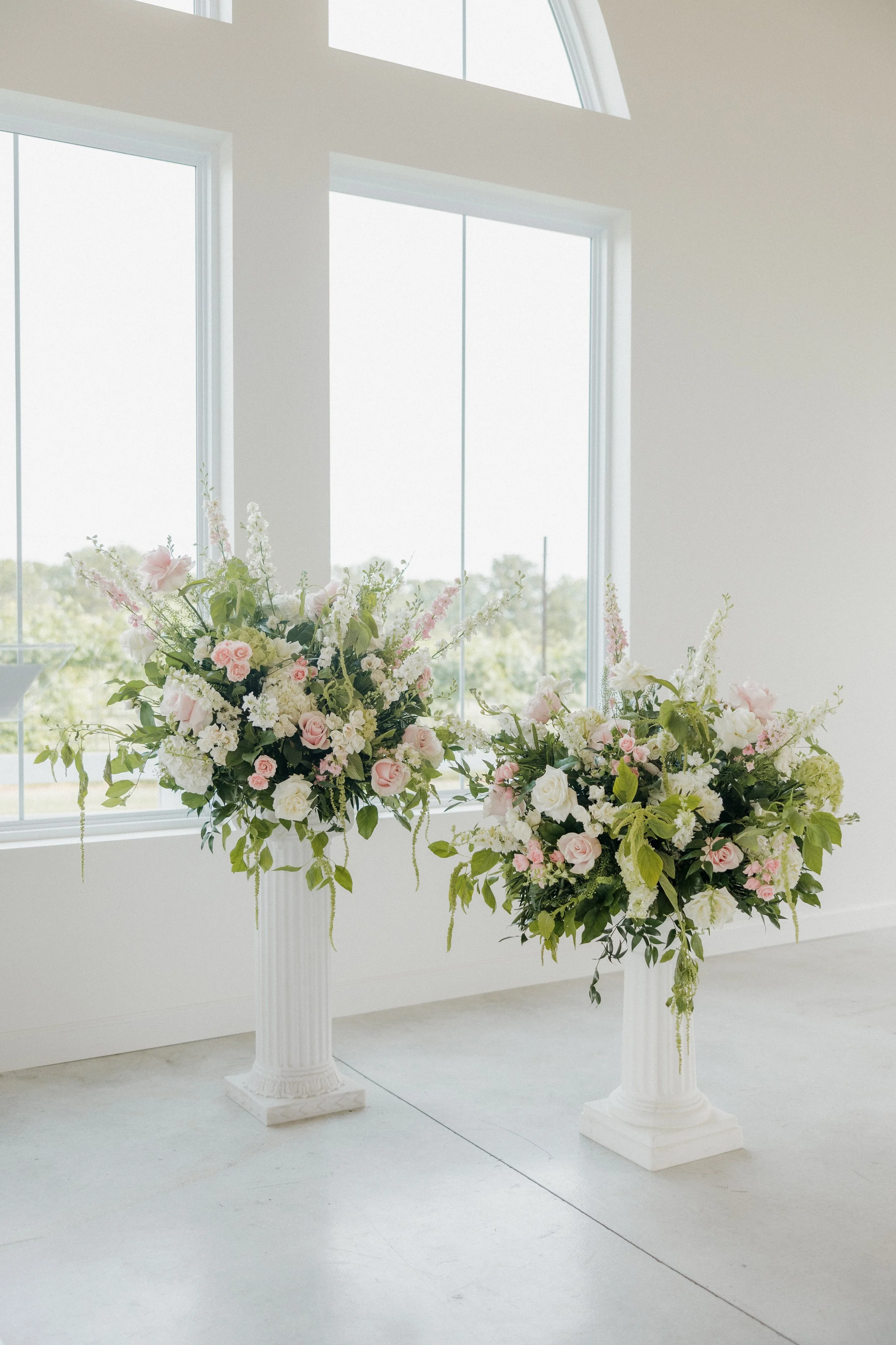 Two large floral arrangements in white classical-style vases with pink and white flowers, set inside a bright, modern room with large windows.