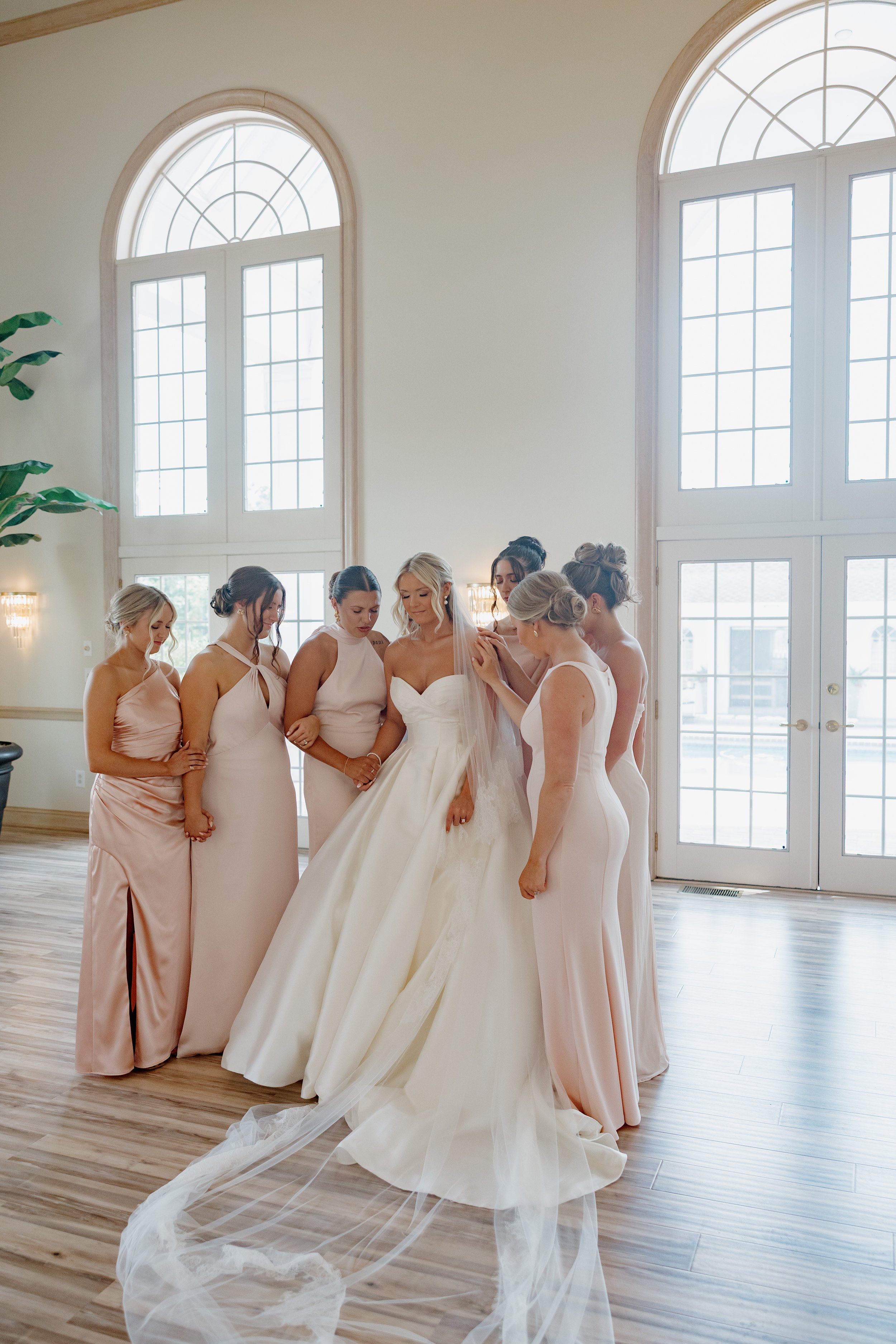 A bride in a white wedding gown surrounded by her bridesmaids in matching pale dresses in a spacious, bright room with large arched windows.