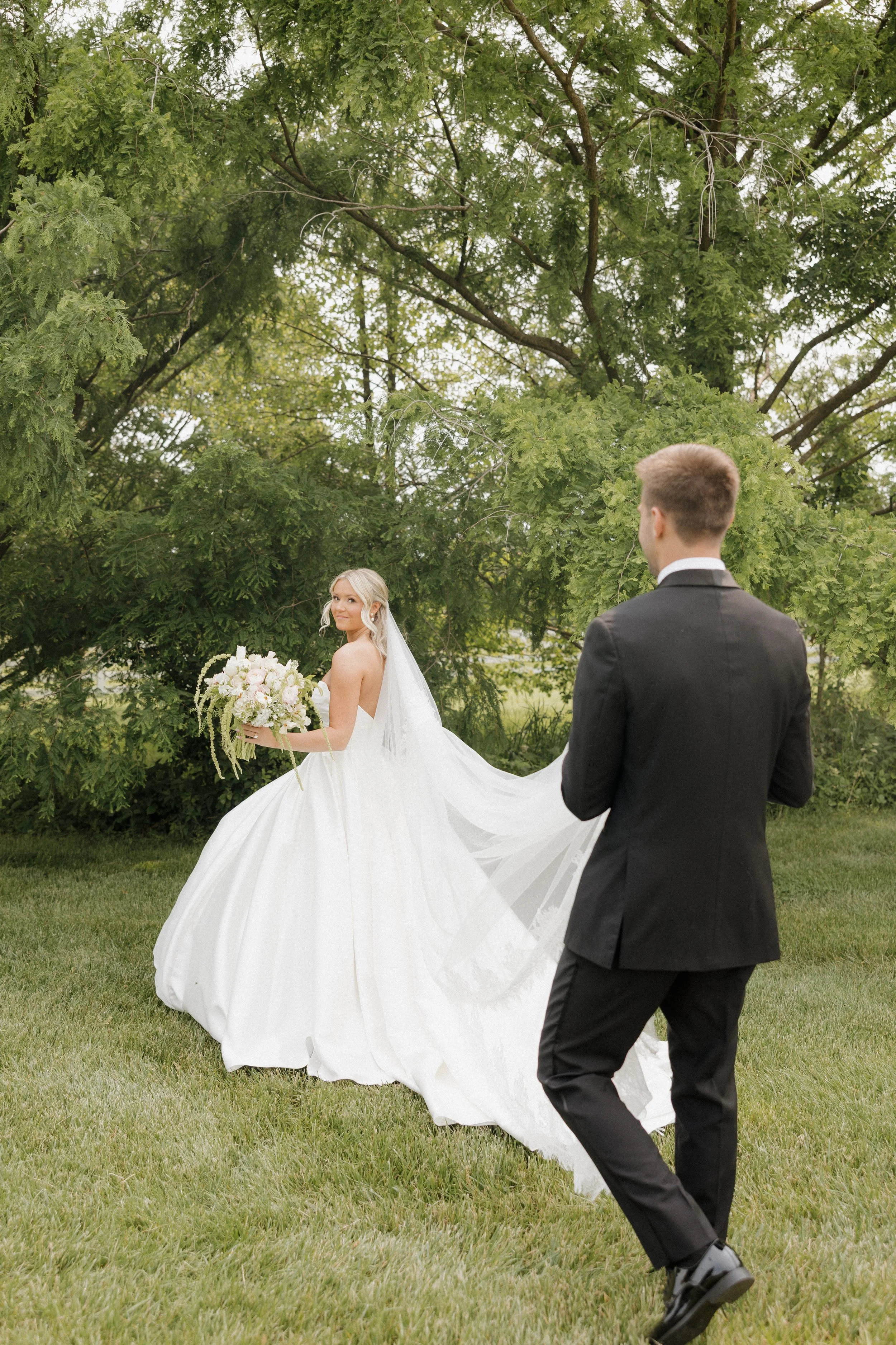 Bride in a white wedding dress holding a bouquet, looking back at groom in a black suit outdoors.