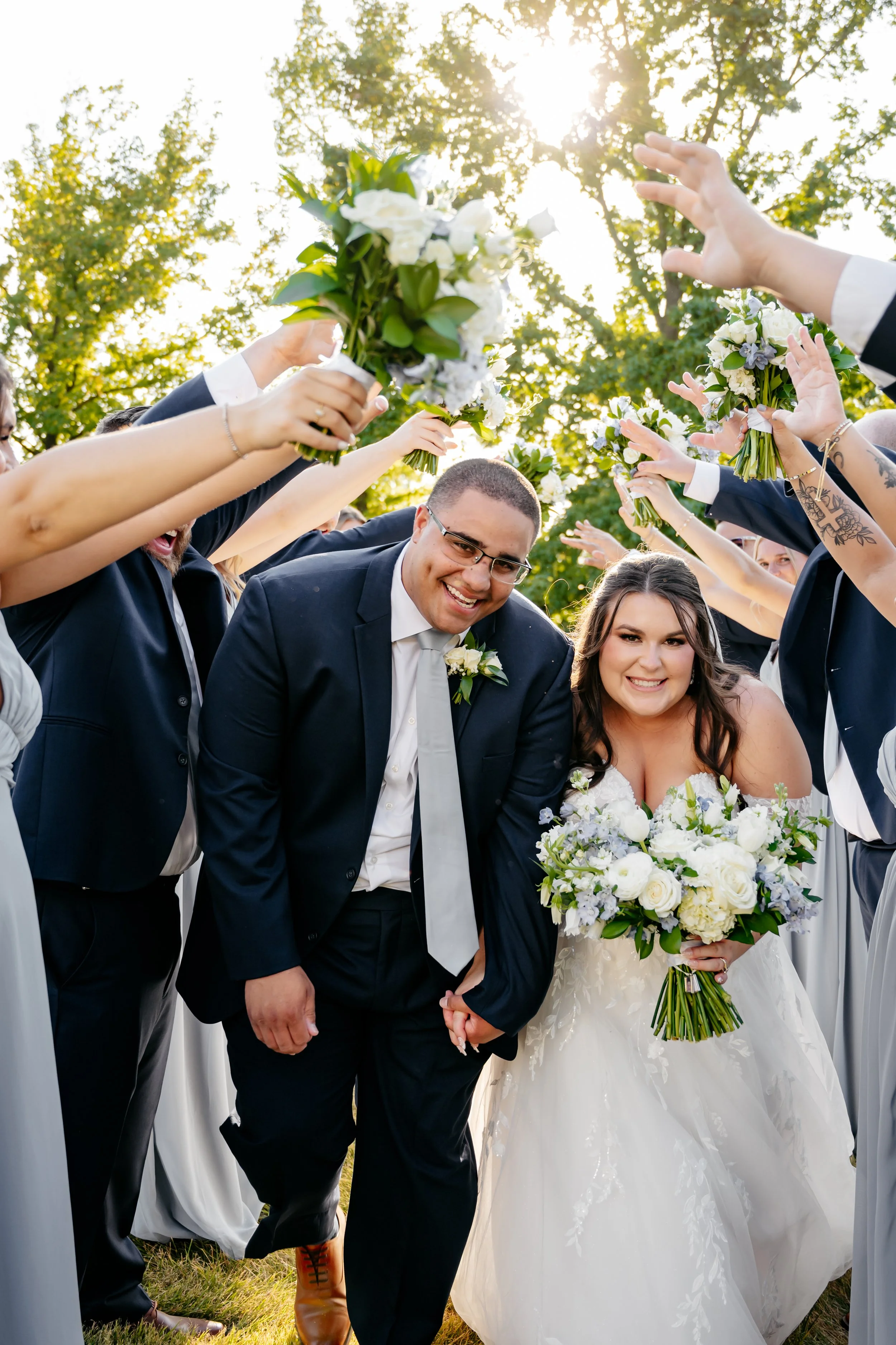 A newlywed couple holding hands and smiling, surrounded by friends and family holding bouquets, during an outdoor wedding celebration in sunlight.