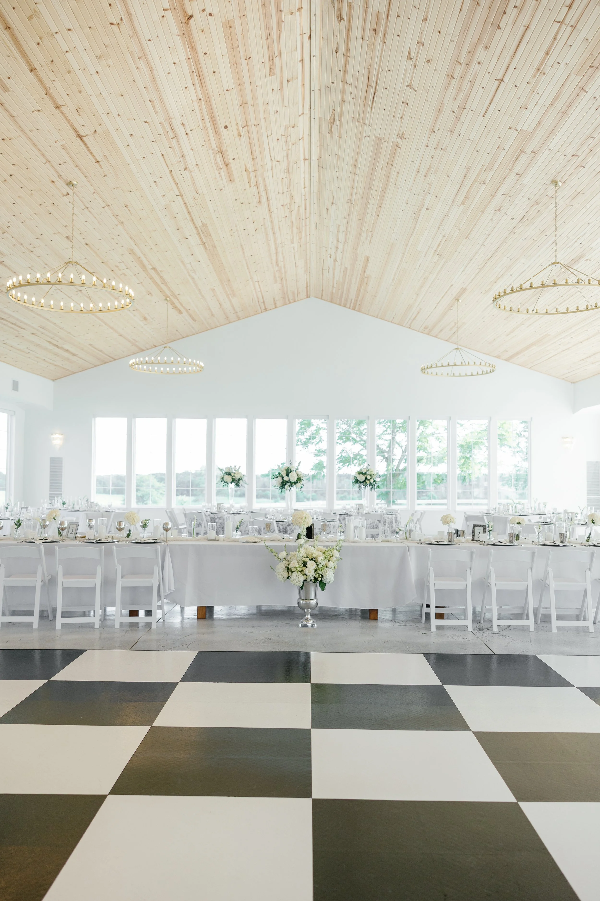 Elegant event hall with a high wooden ceiling, circular chandeliers, large windows, and a long table set with white tablecloths, floral centerpieces, and tableware. Black and white checkered floor in the foreground.