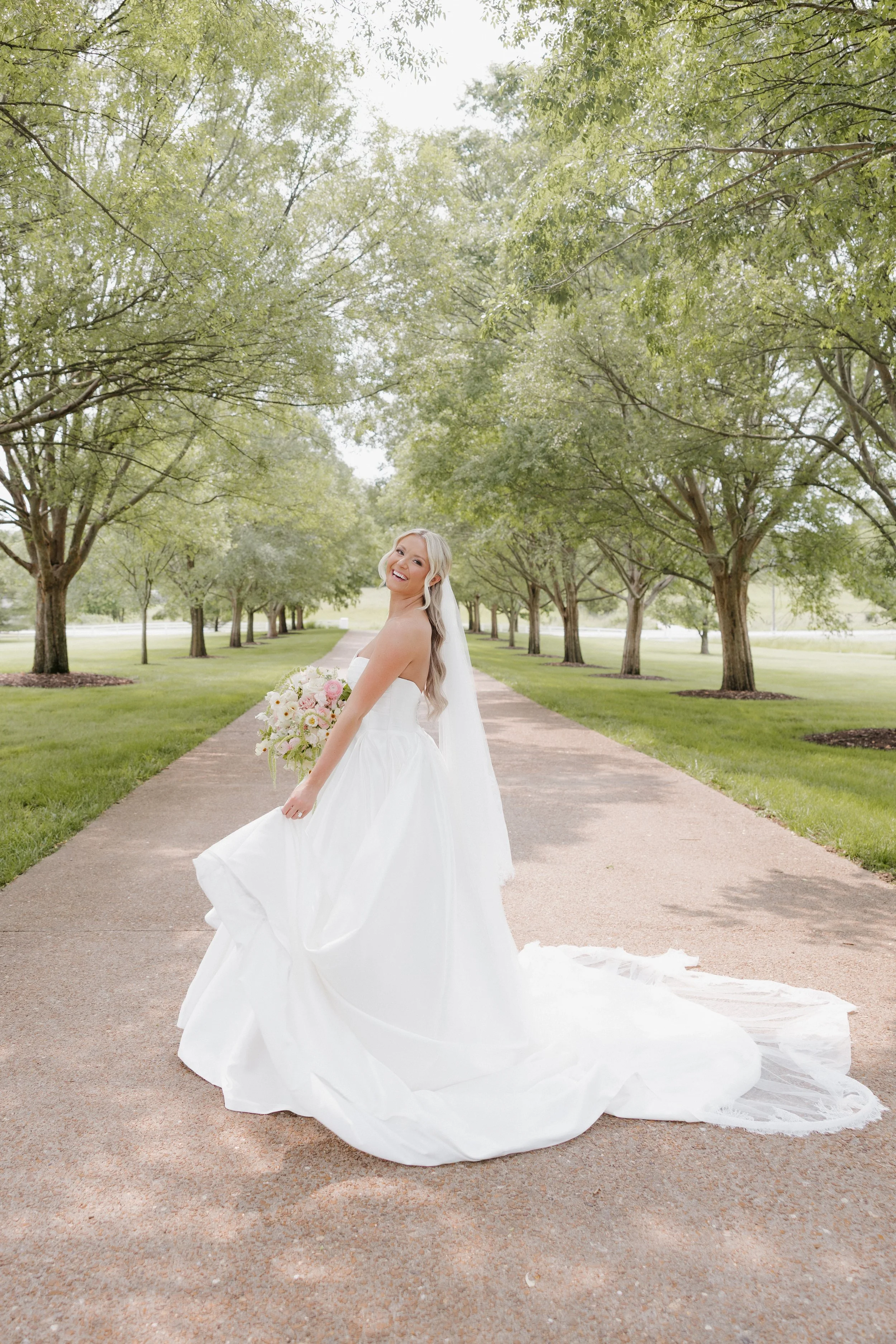 A joyful bride in a white wedding gown holding a bouquet of pink and white flowers, standing on a tree-lined pathway in a park.