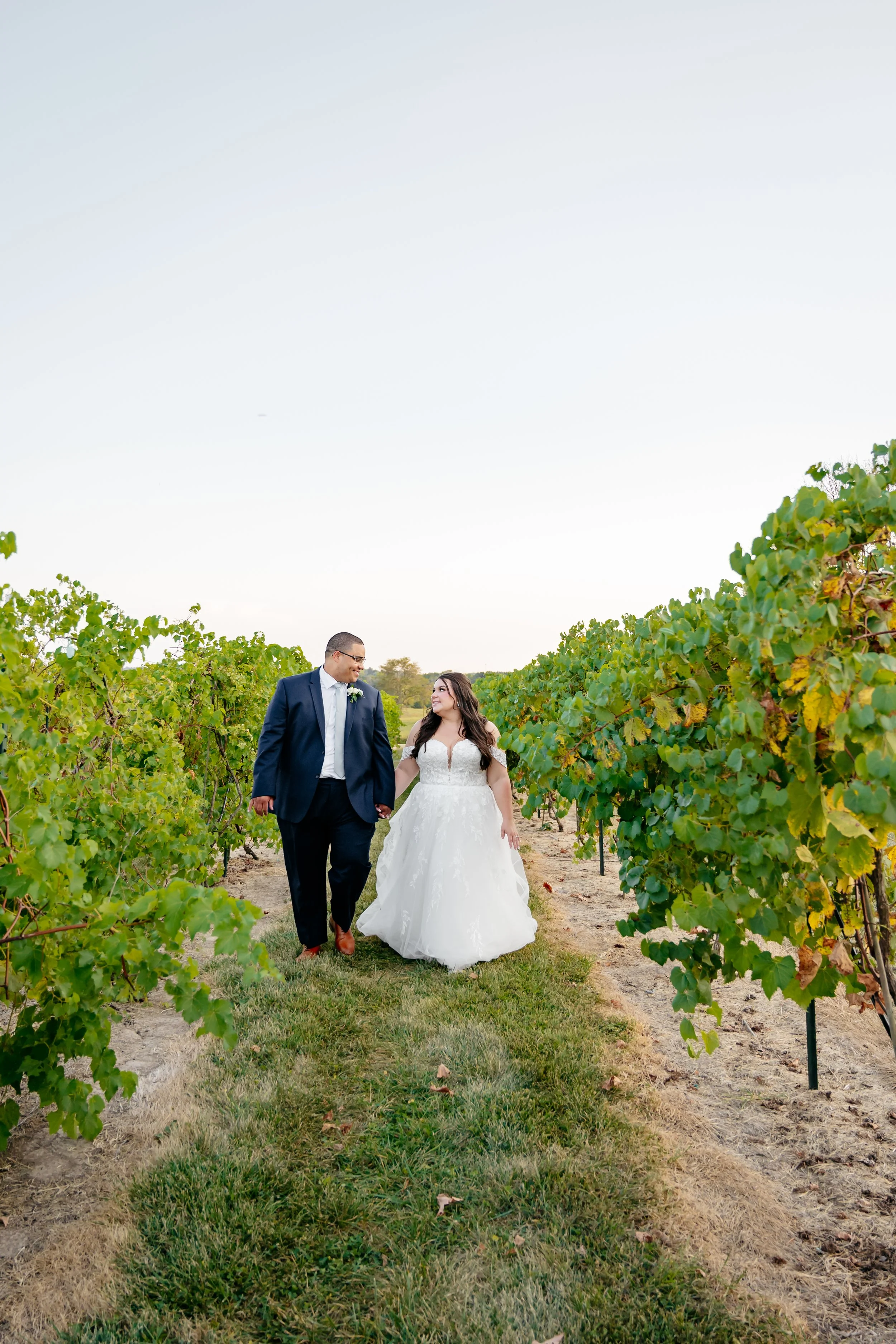 A bride and groom walking hand in hand through a vineyard, smiling at each other, as part of a wedding photoshoot.