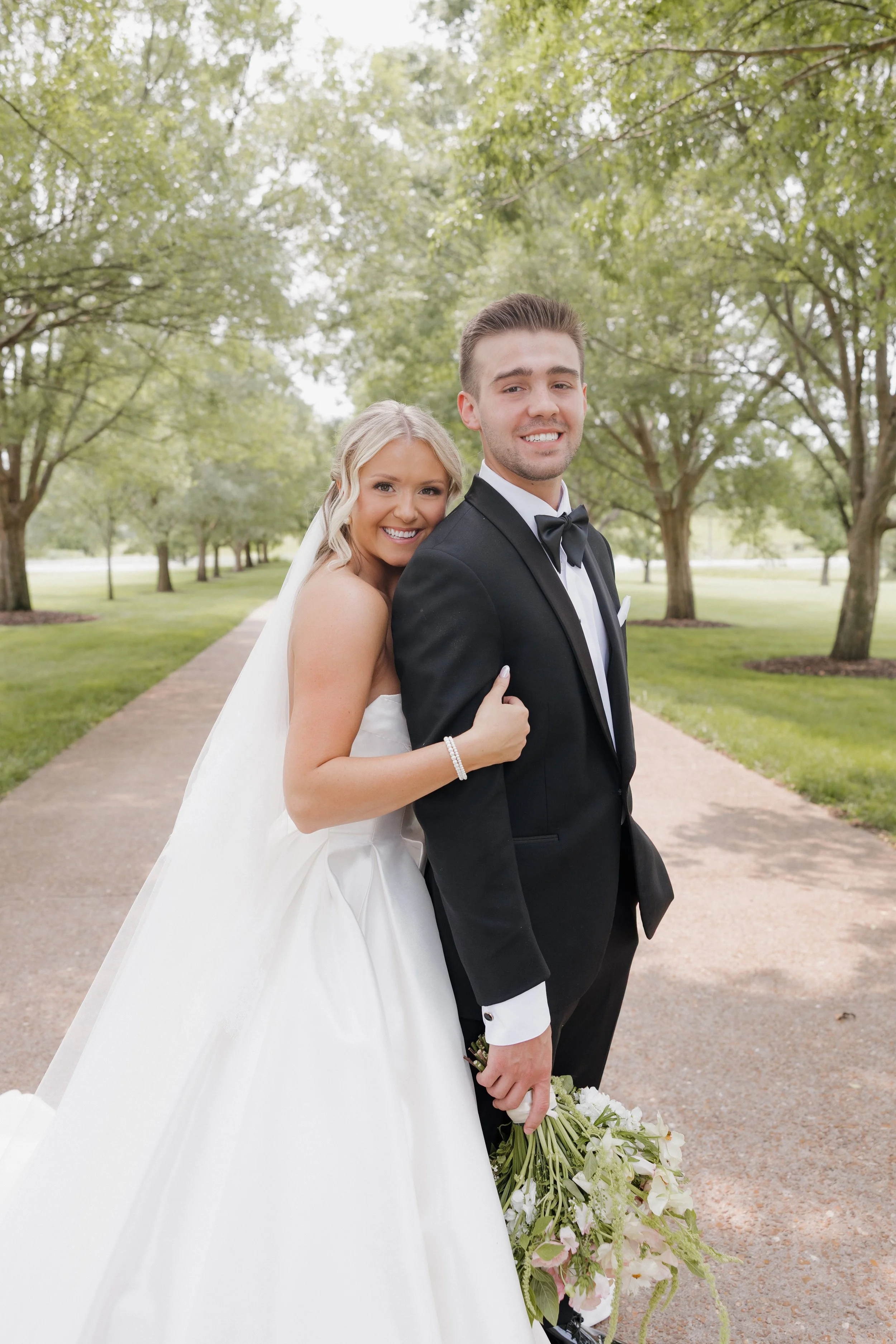 A bride and groom standing closely together outdoors on a pathway surrounded by green trees, smiling at the camera. The bride is in a white wedding gown holding a bouquet of flowers, and the groom is in a black tuxedo with a bow tie.