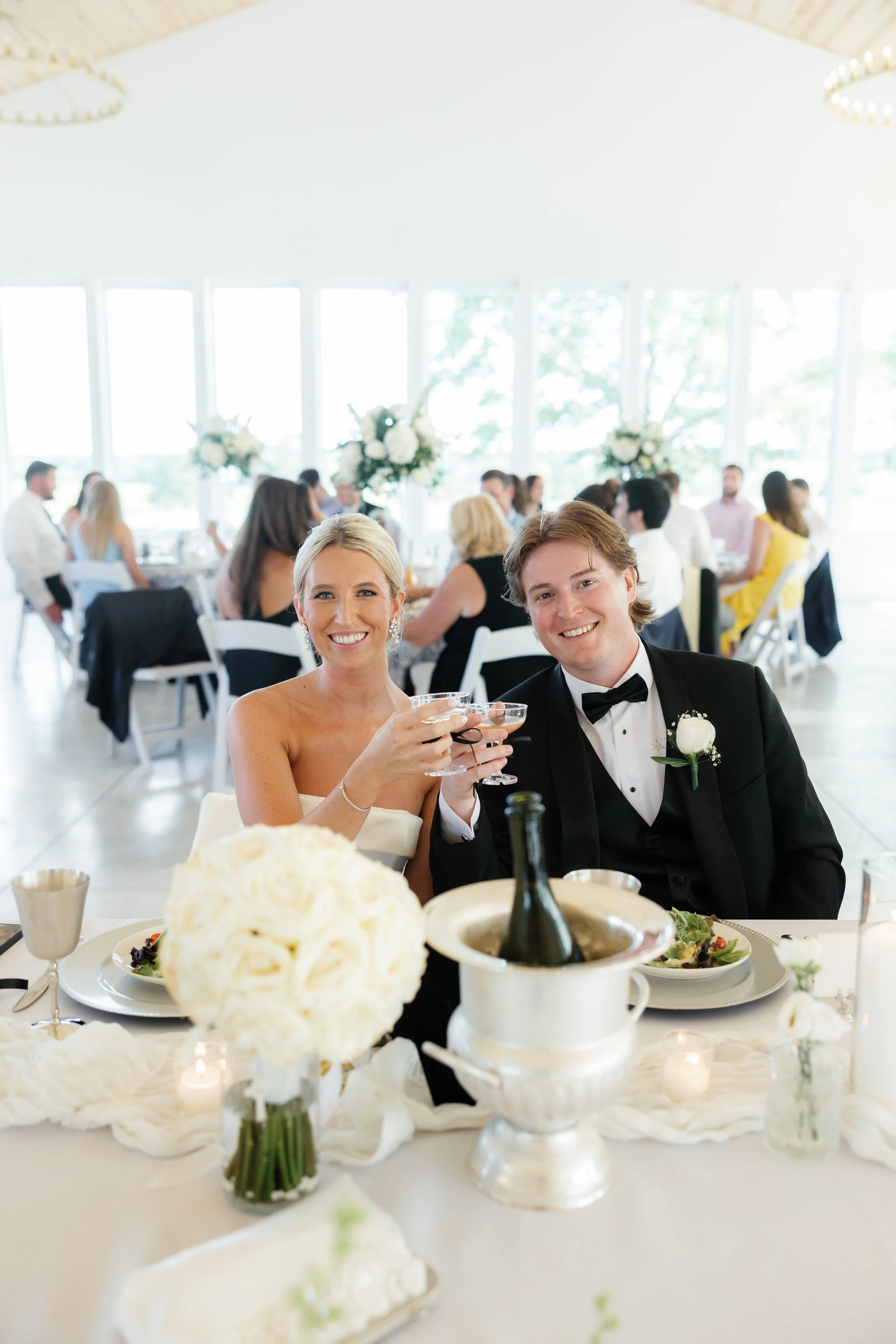A bride and groom sit at a wedding reception table, raising glasses in a toast, smiling. The table is decorated with white flowers, candles, and a champagne bottle in an ice bucket. The reception hall has large windows, allowing natural light to fill