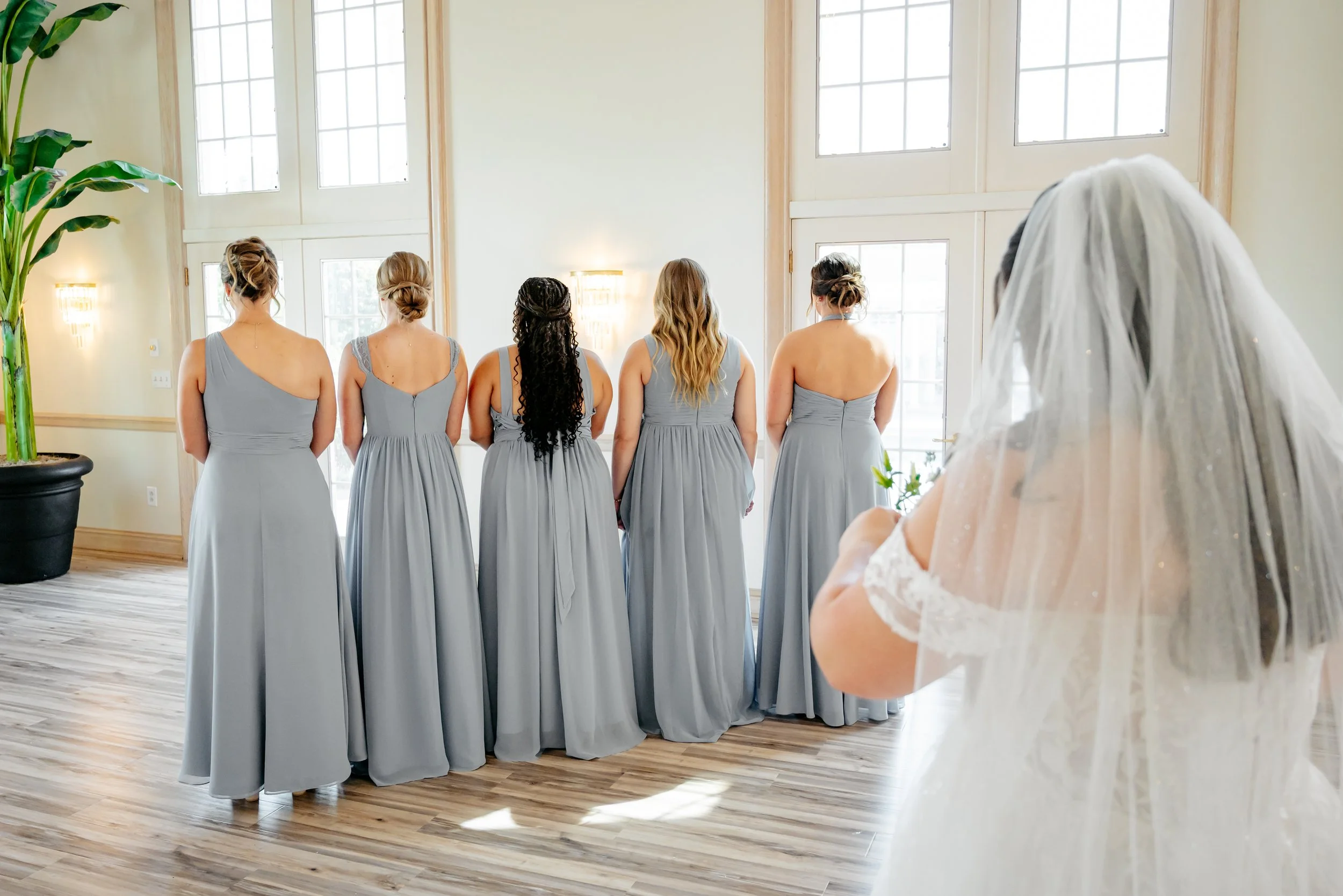 Bride in a wedding dress with veil watching five bridesmaids in gray dresses standing at the window in a bright room with large windows and wooden floors.