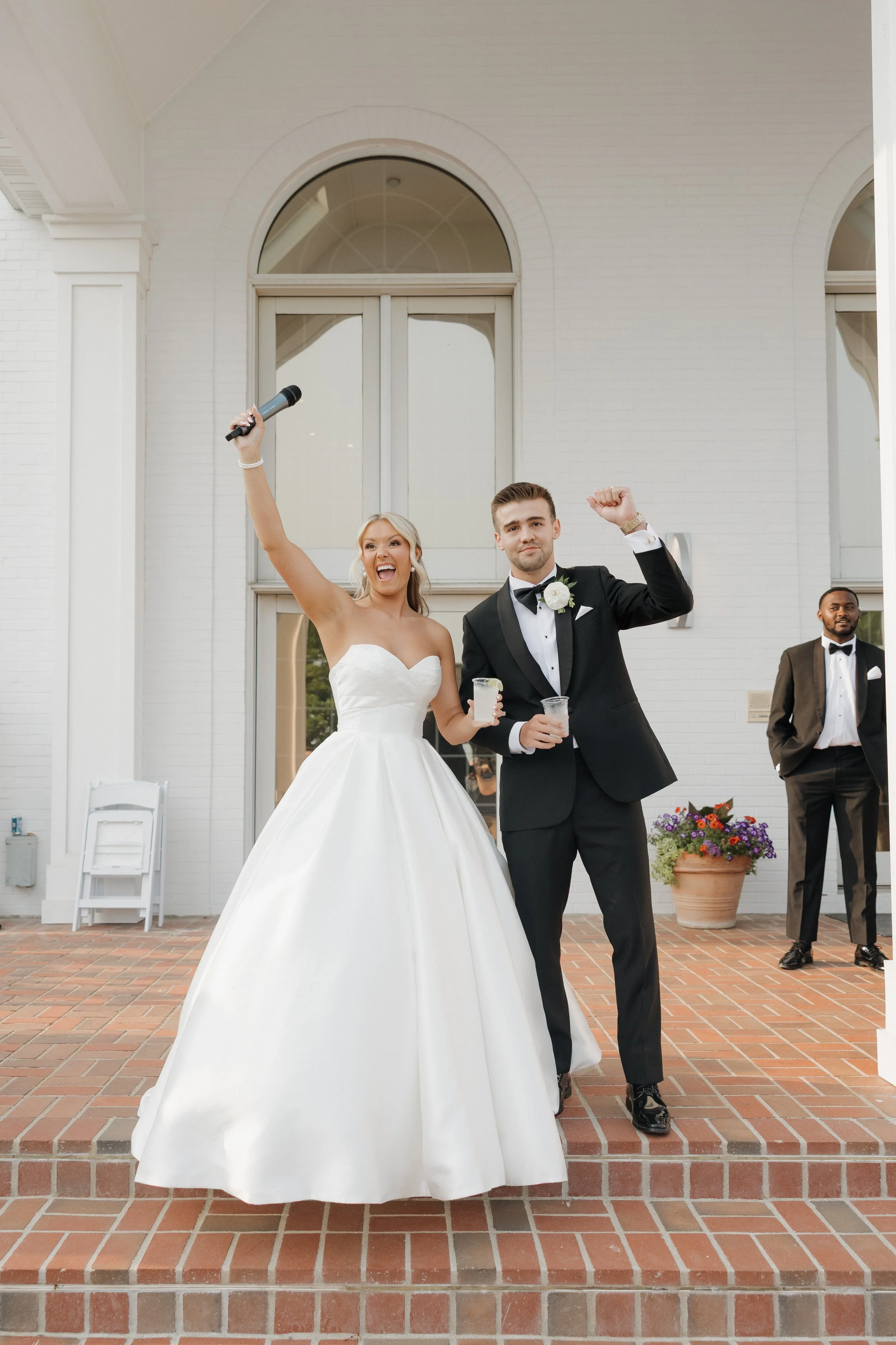 Bride in a white wedding gown and groom in a black tuxedo celebrating with drinks outside a building. The bride is holding a microphone and smiling, while the groom is raising his fist in celebration.