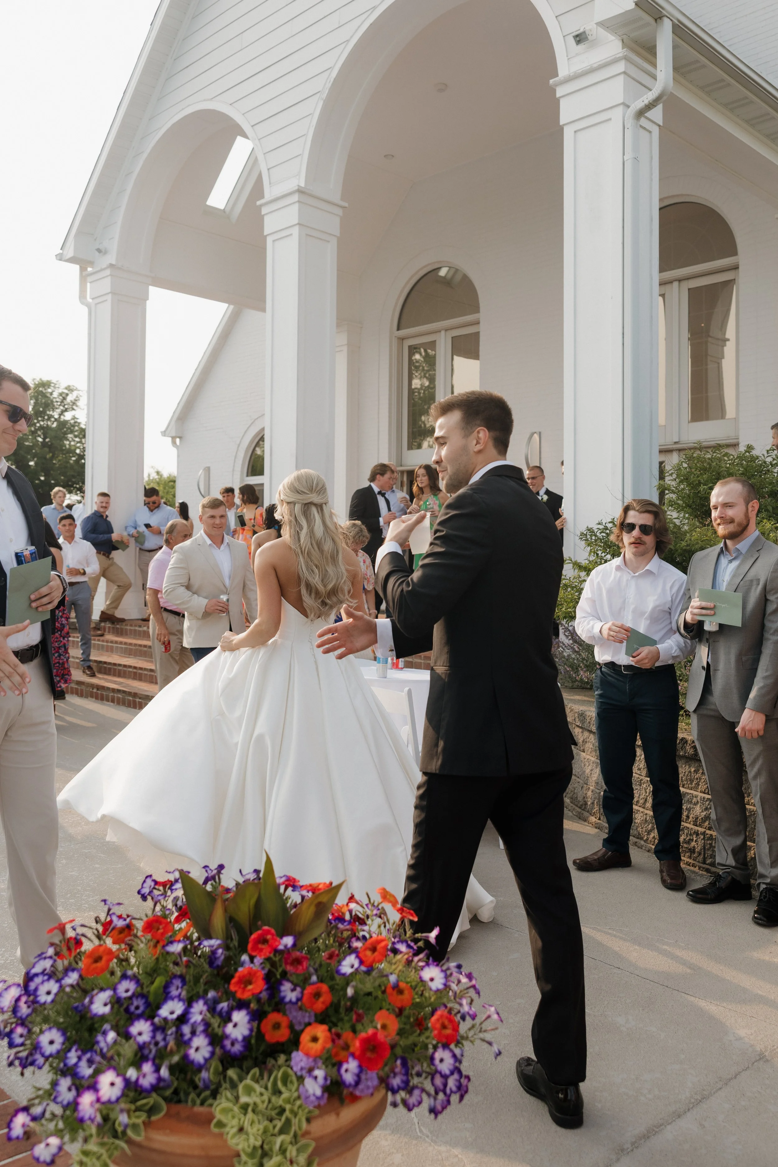 Wedding guests gather outside a white church, with a bride in a white gown and a groom in a black tuxedo, surrounded by friends, some holding programs, as they celebrate the wedding ceremony.