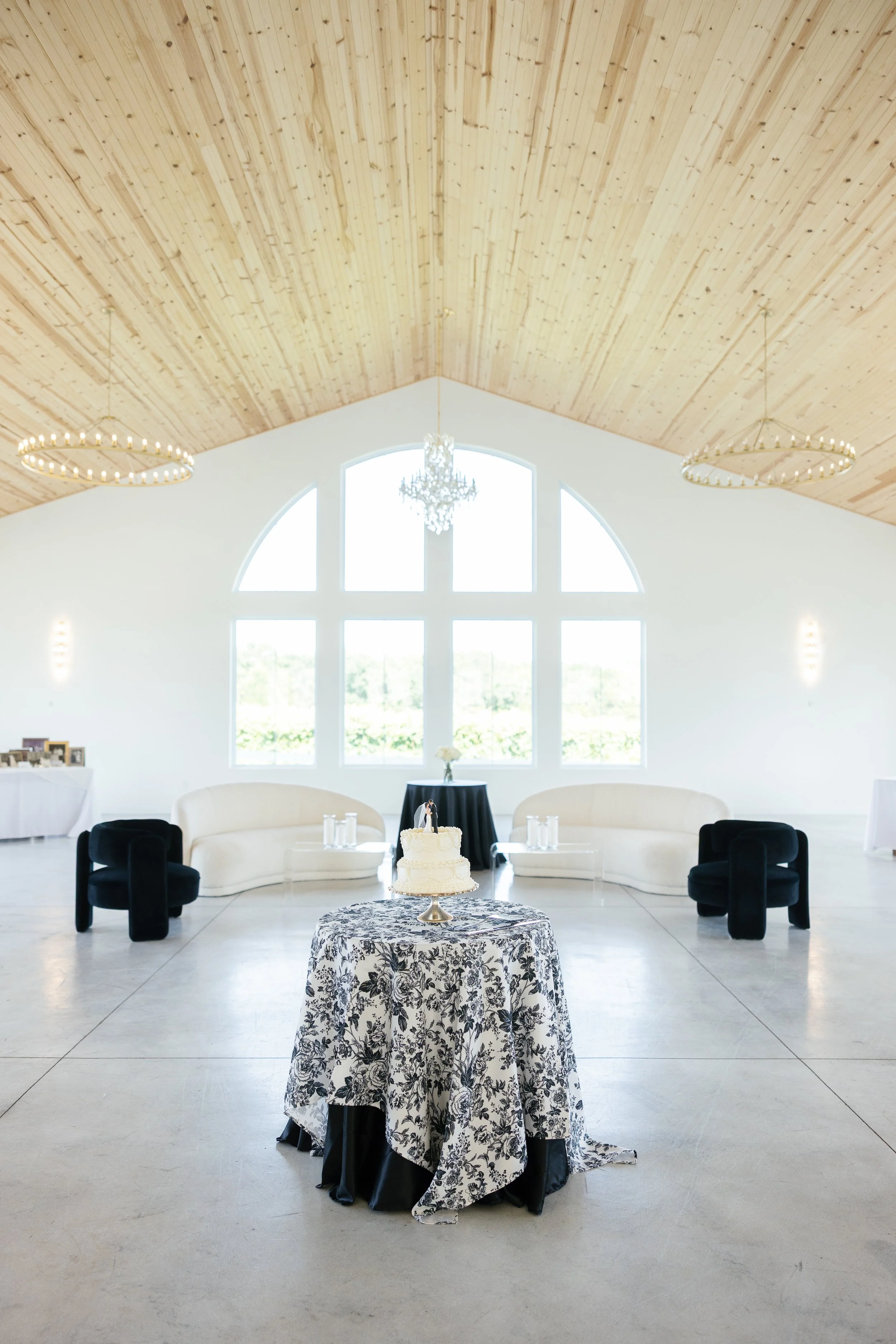 White wedding reception setup with a tiered cake on a round table with black and white floral tablecloth. Behind the cake are two white curved sofas, with a black table and a flower arrangement in the middle. Large arched windows and chandeliers are 