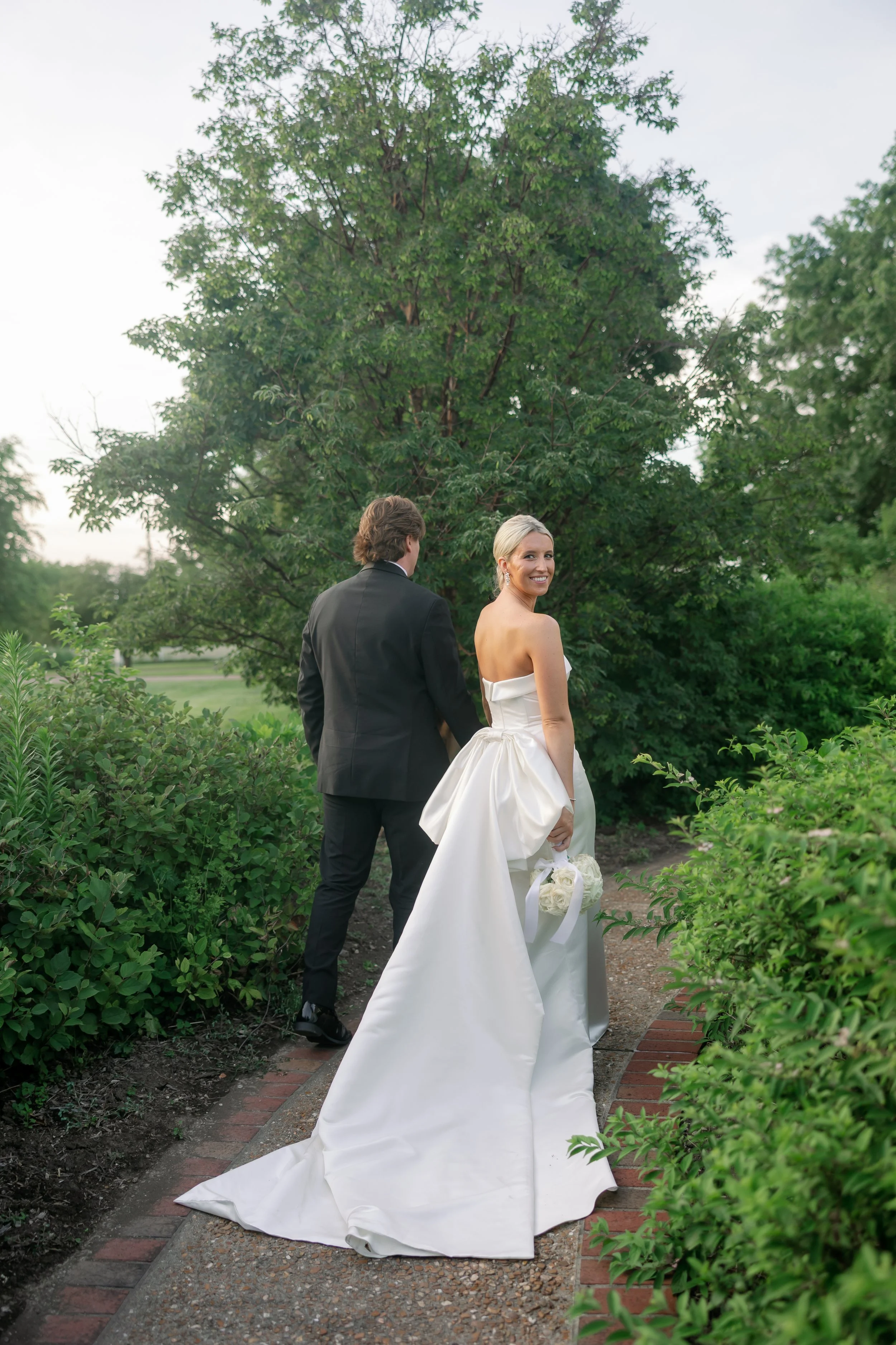 A bride and groom standing on a garden path, with the bride smiling at the camera and holding a bouquet of white flowers, while the groom faces away, surrounded by greenery and trees.
