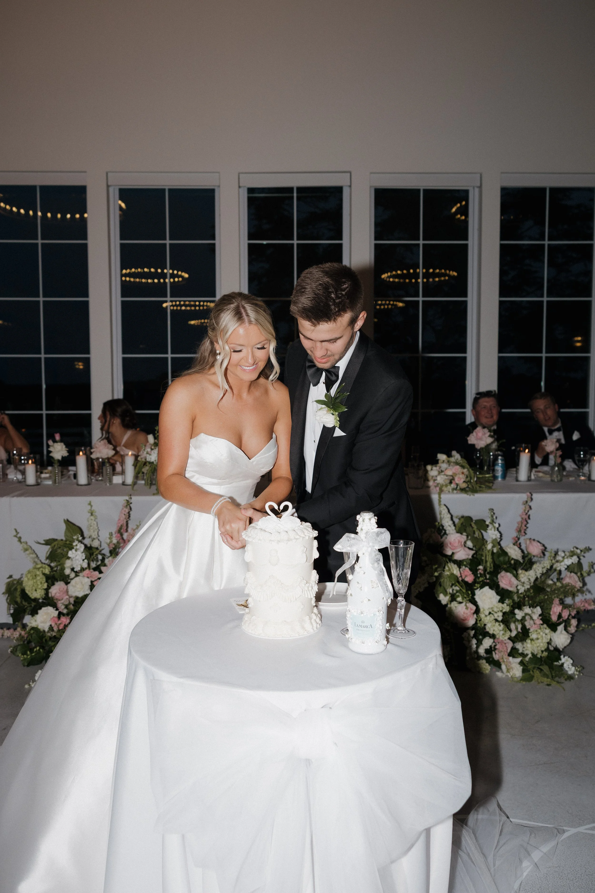 Bride and groom cutting their wedding cake at a wedding reception with floral decorations in the background.