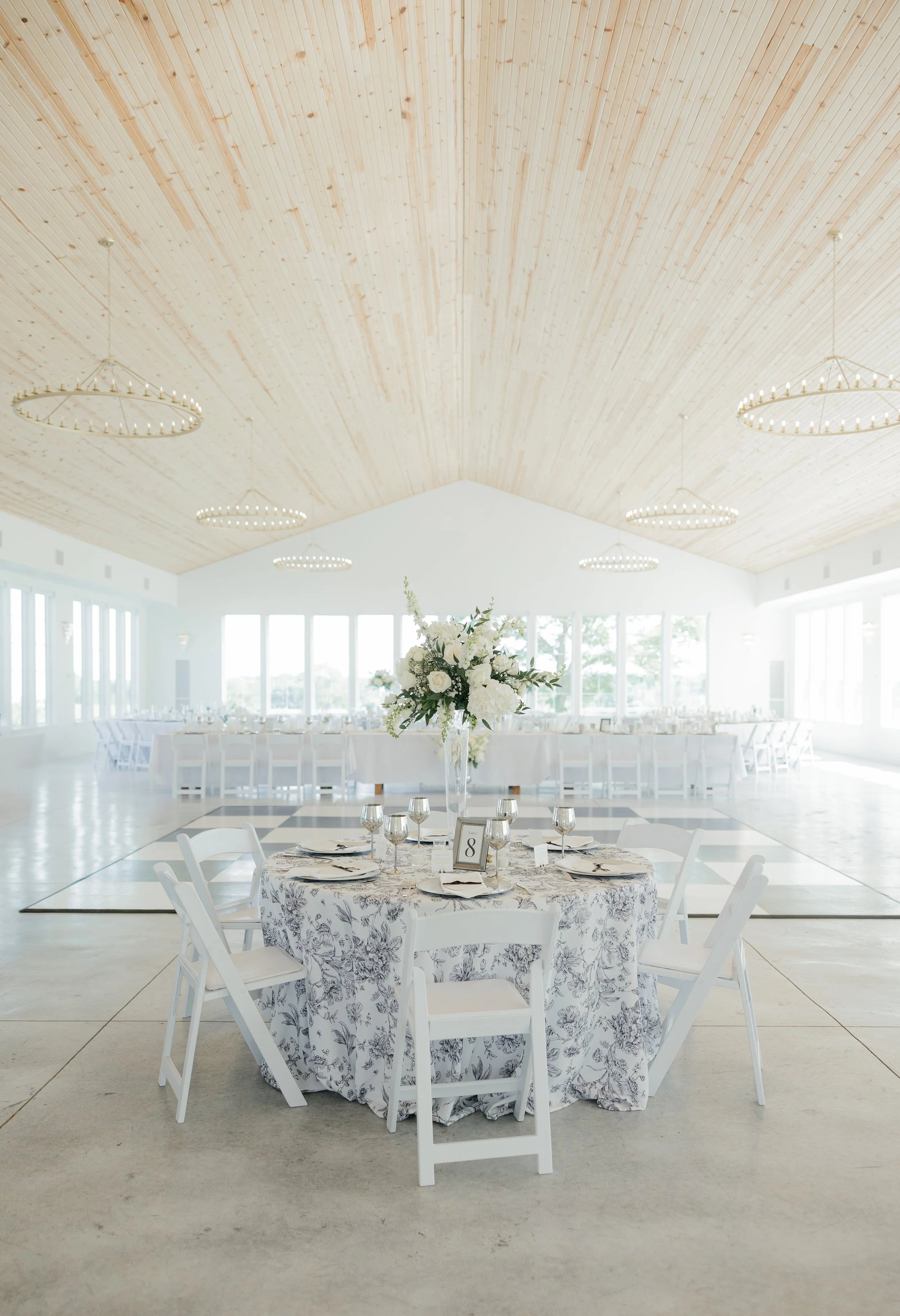Elegant indoor wedding reception with a round table covered in a floral tablecloth, set with plates, wine glasses, and utensils, featuring a tall floral centerpiece with white flowers, and surrounded by white chairs. In the background, there are addi