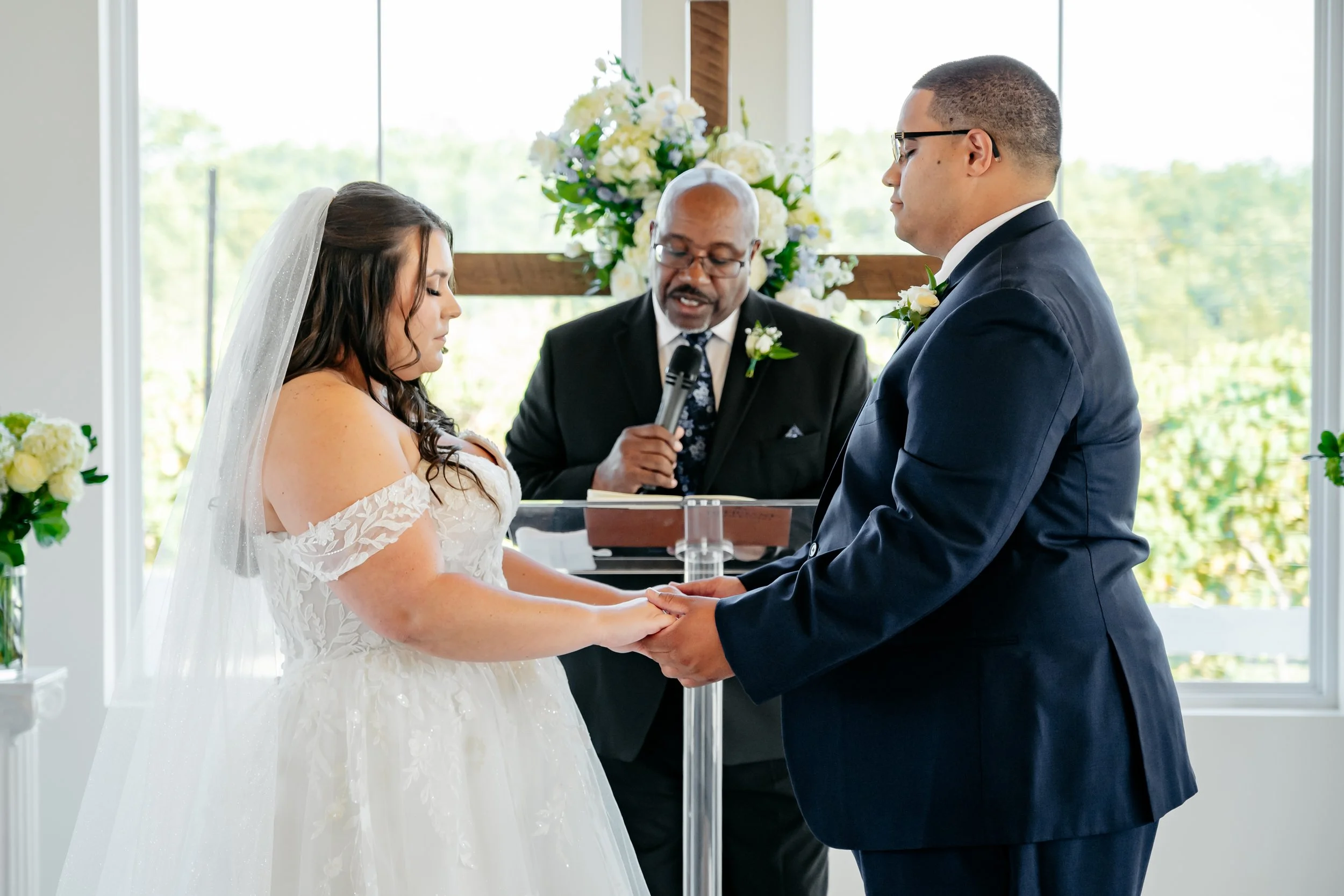 A bride and groom holding hands during their wedding ceremony, with an officiant reading from a book, inside a bright room with large windows and floral arrangements.