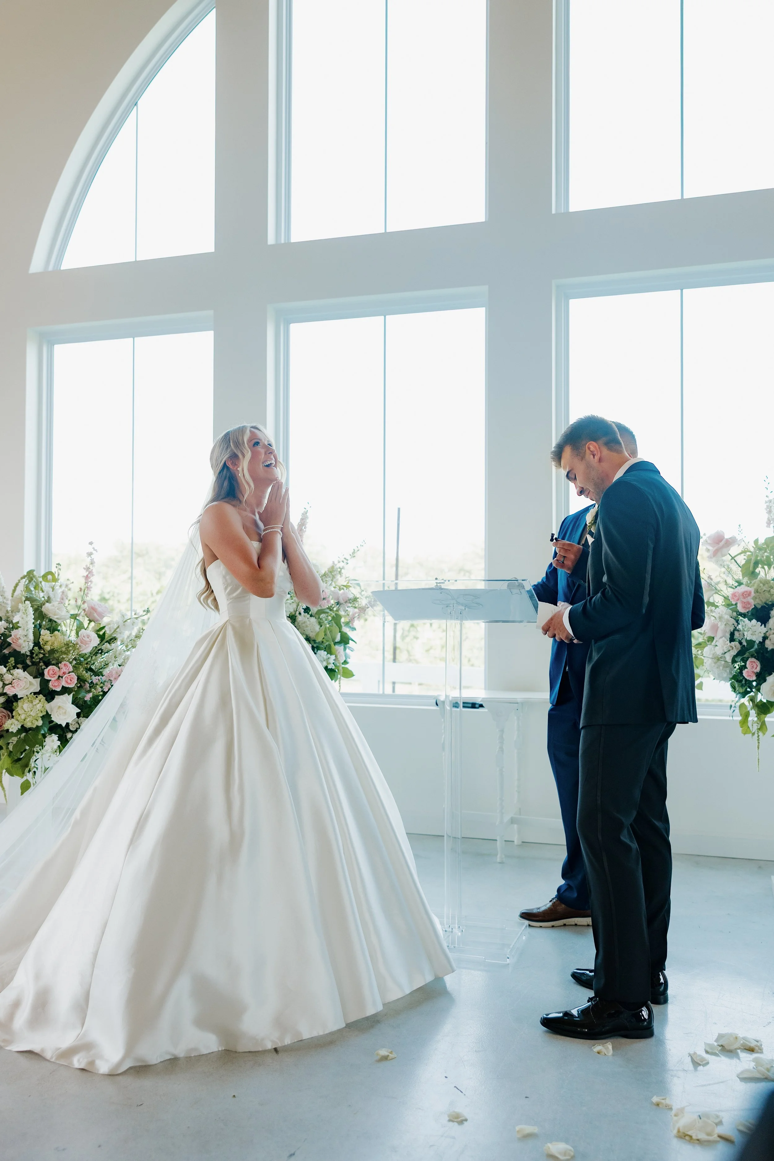 A bride in a white wedding dress with a full skirt and a veil is smiling happily at her wedding ceremony. Two groomsmen in dark suits are standing beside her, possibly reading vows or giving speeches. The ceremony is taking place in a bright room wit