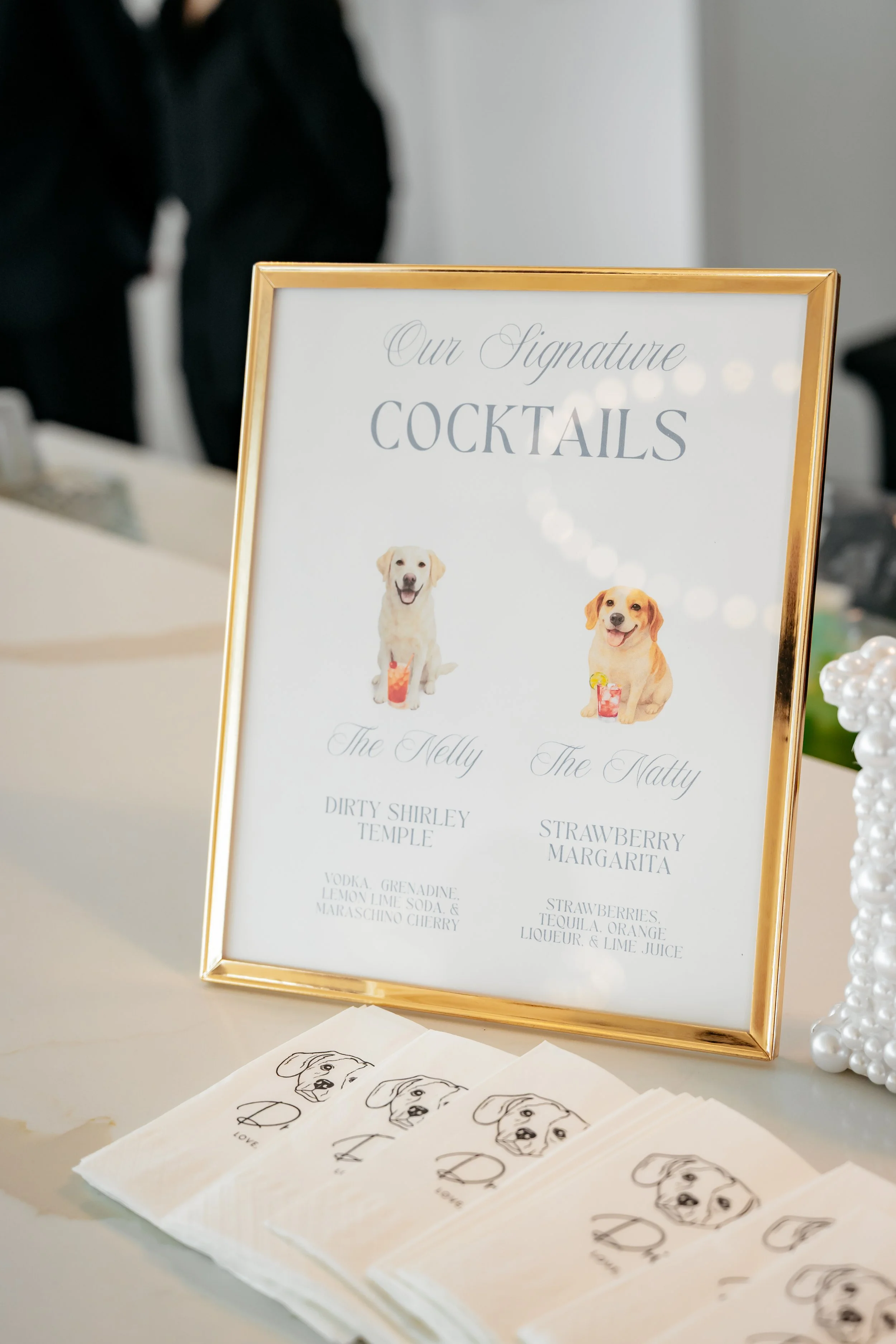 Sign with dog illustrations and cocktail menu at an event, titled "Our Signature Cocktails" with options "The Nelly" and "The Natty."