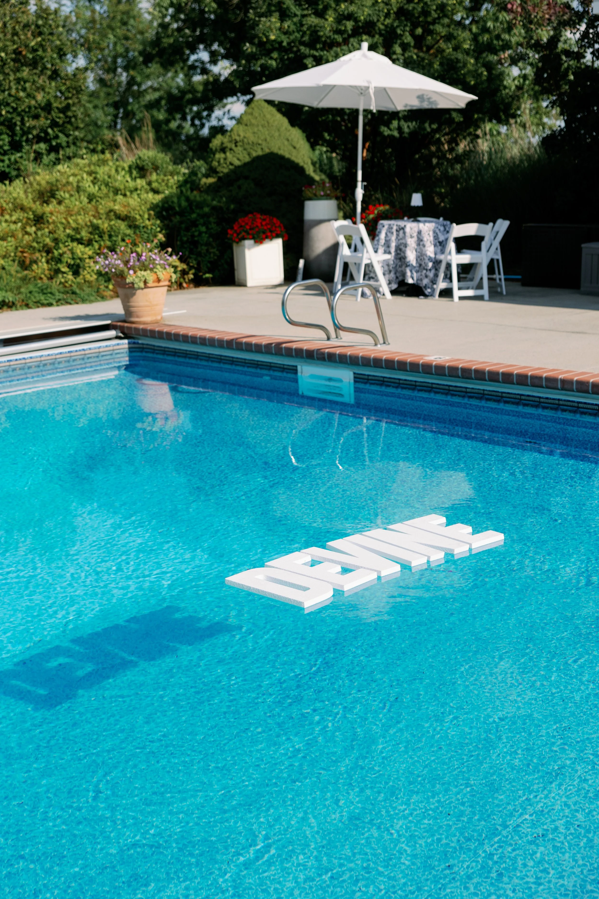 Empty outdoor swimming pool with a floating "SKIP" sign and a patio area in the background, featuring a table with a tablecloth, chairs, a large umbrella, and potted plants.