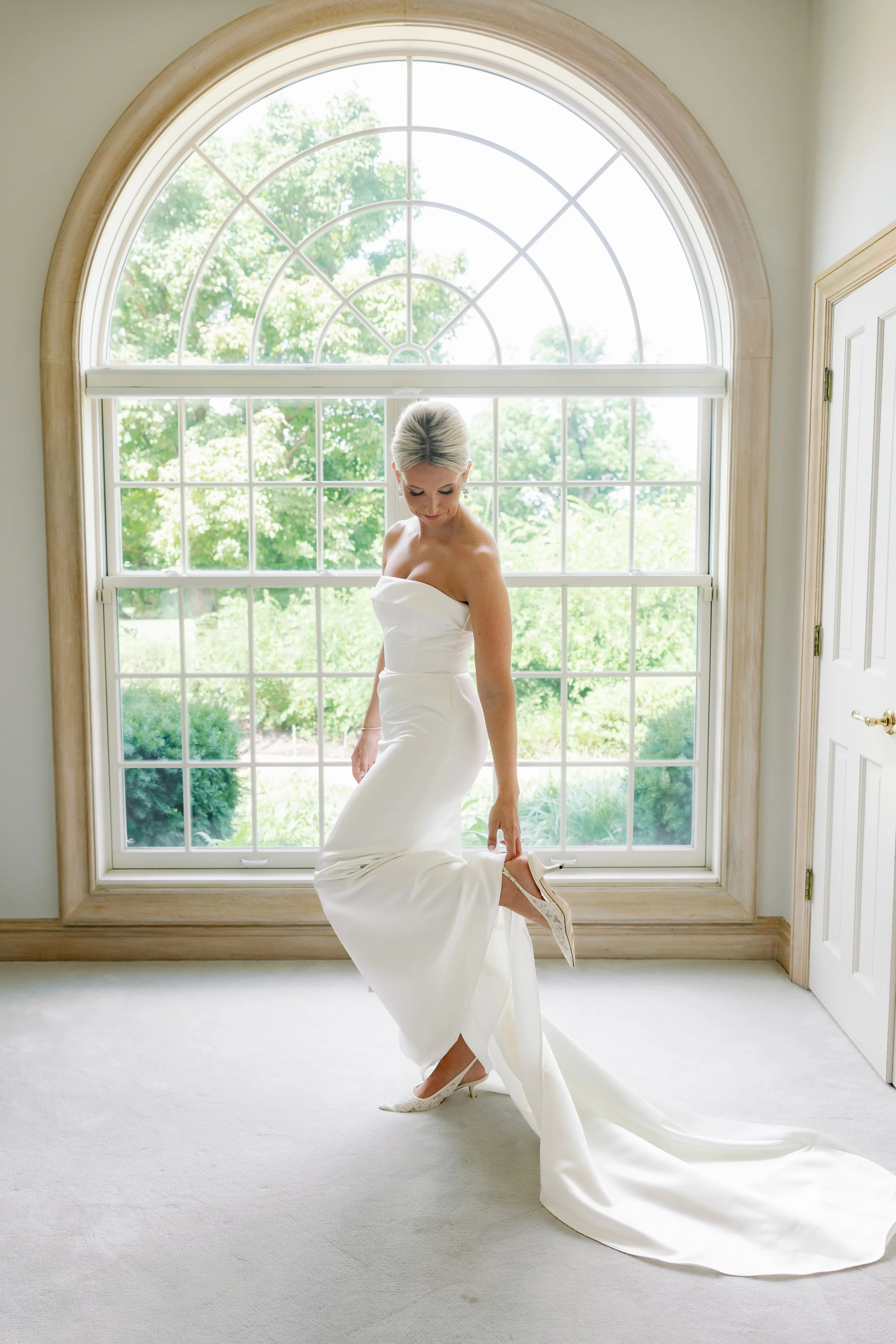 Bride in a strapless white wedding gown adjusting her white high heel shoe in front of a large arched window with greenery outside.