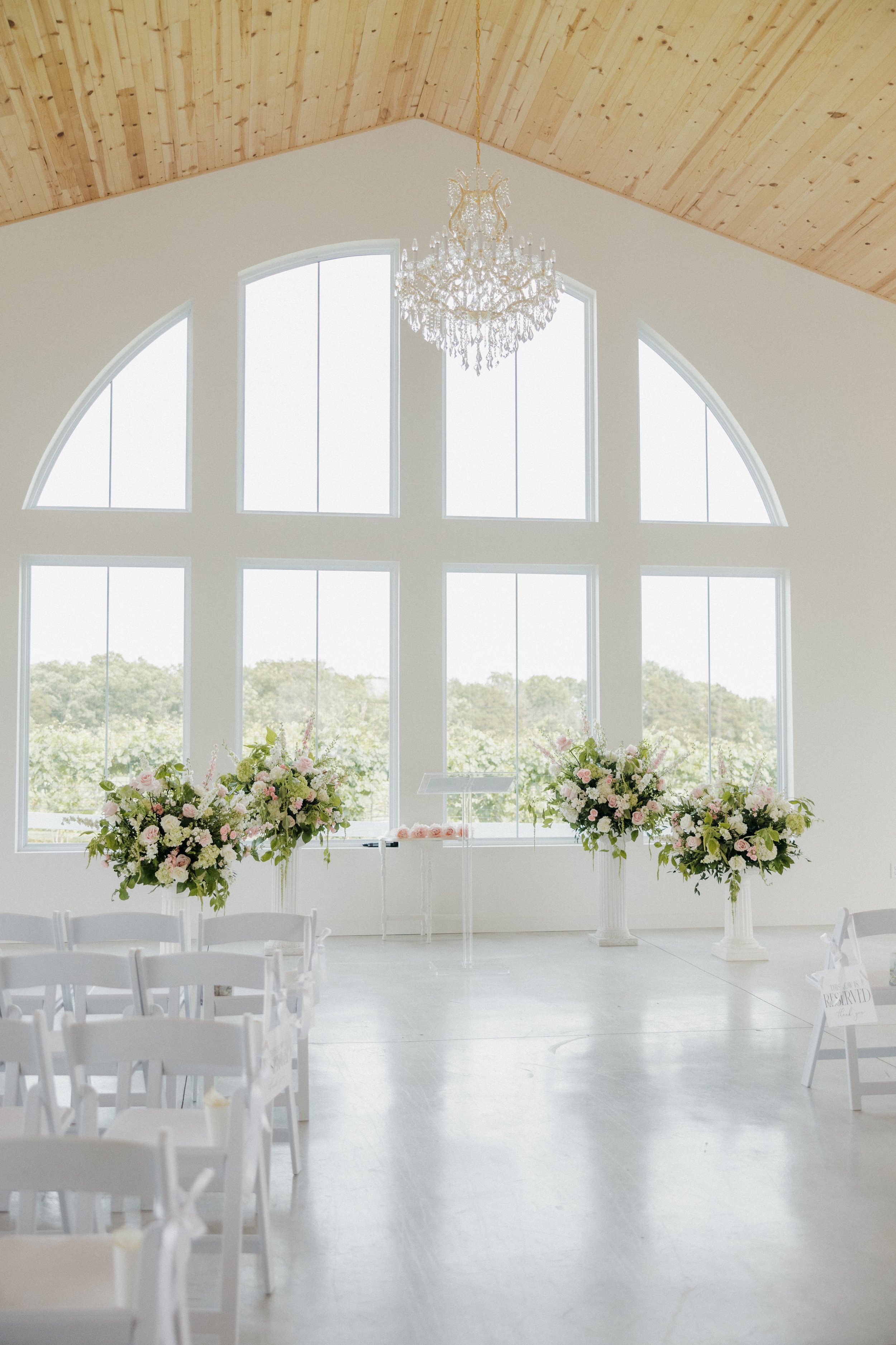Empty wedding ceremony space with large arched windows, floral arrangements, and a chandelier.