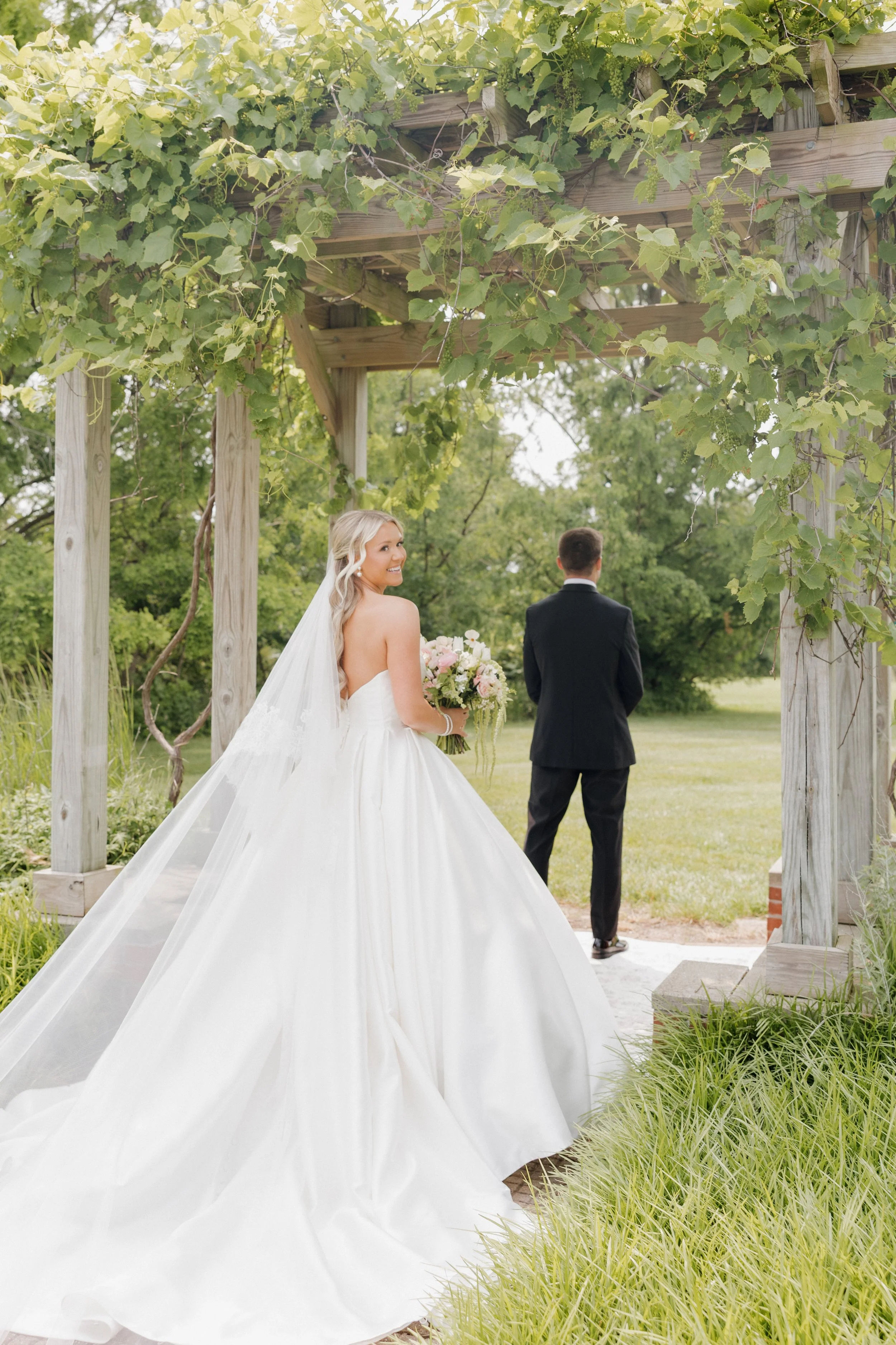 Bride in a white wedding gown holding a bouquet and smiling, standing under a wooden pergola covered with green vines, with the groom in a black suit facing away in the background.