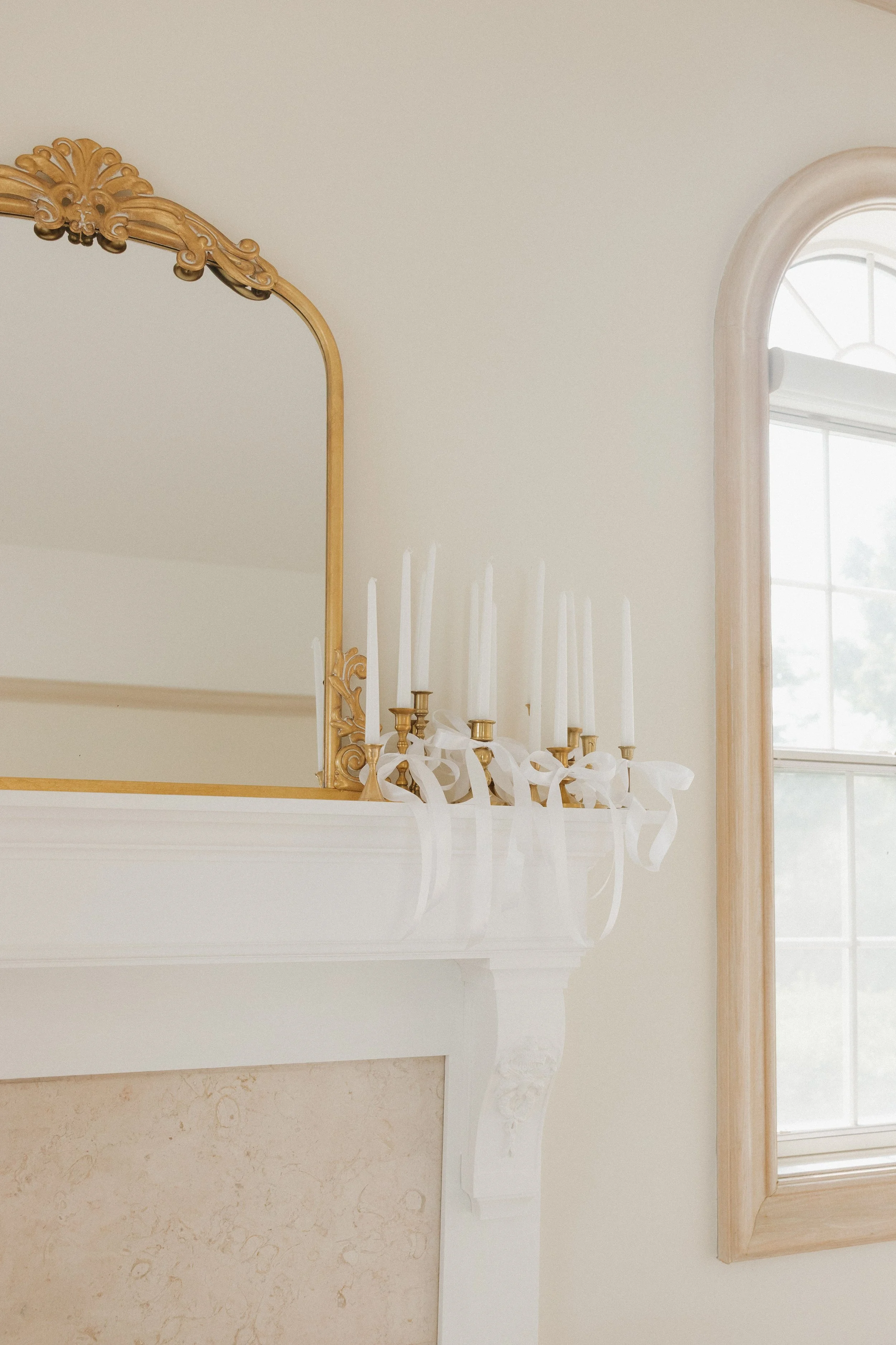 A decorated white fireplace mantel with white candles tied with white ribbons, a gold and mirror above it, and a window next to it with a wooden frame.