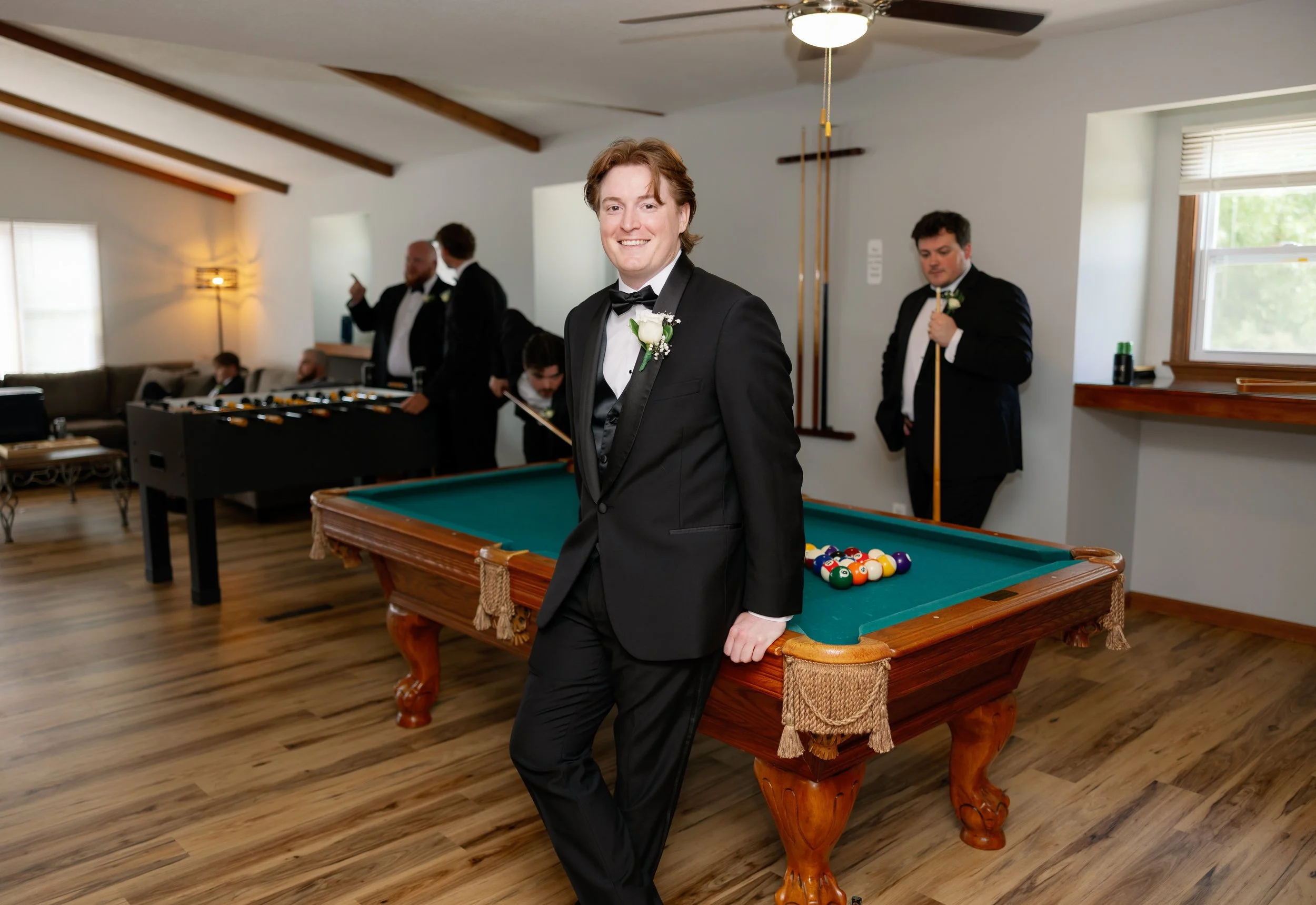 A young man in a black tuxedo with a white shirt and bow tie, smiling and standing with one hand on a billiards table with pool balls, in a room with other men in tuxedos.