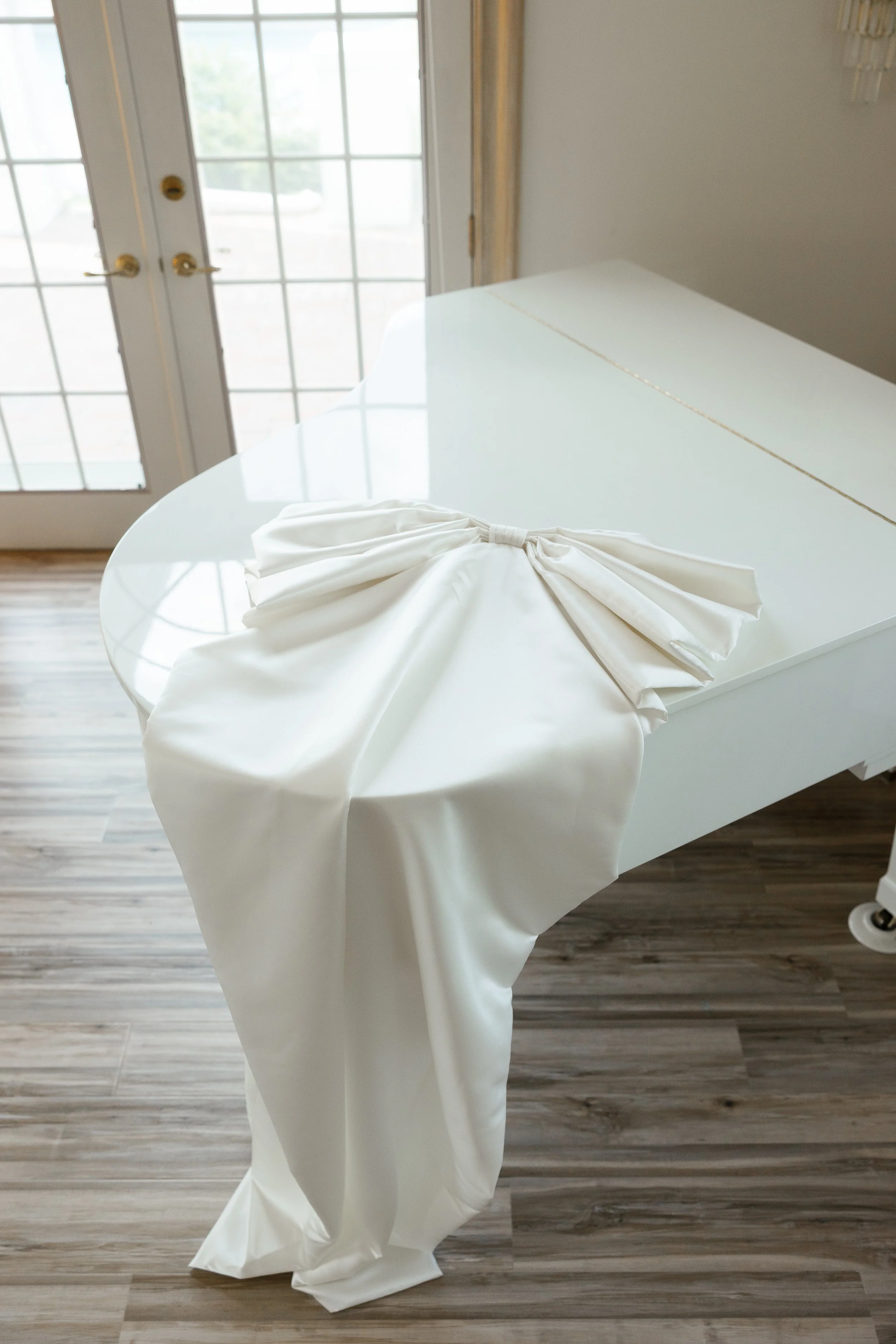 A white grand piano with a white satin cloth draped over its edge, placed near glass double doors with gold handles.