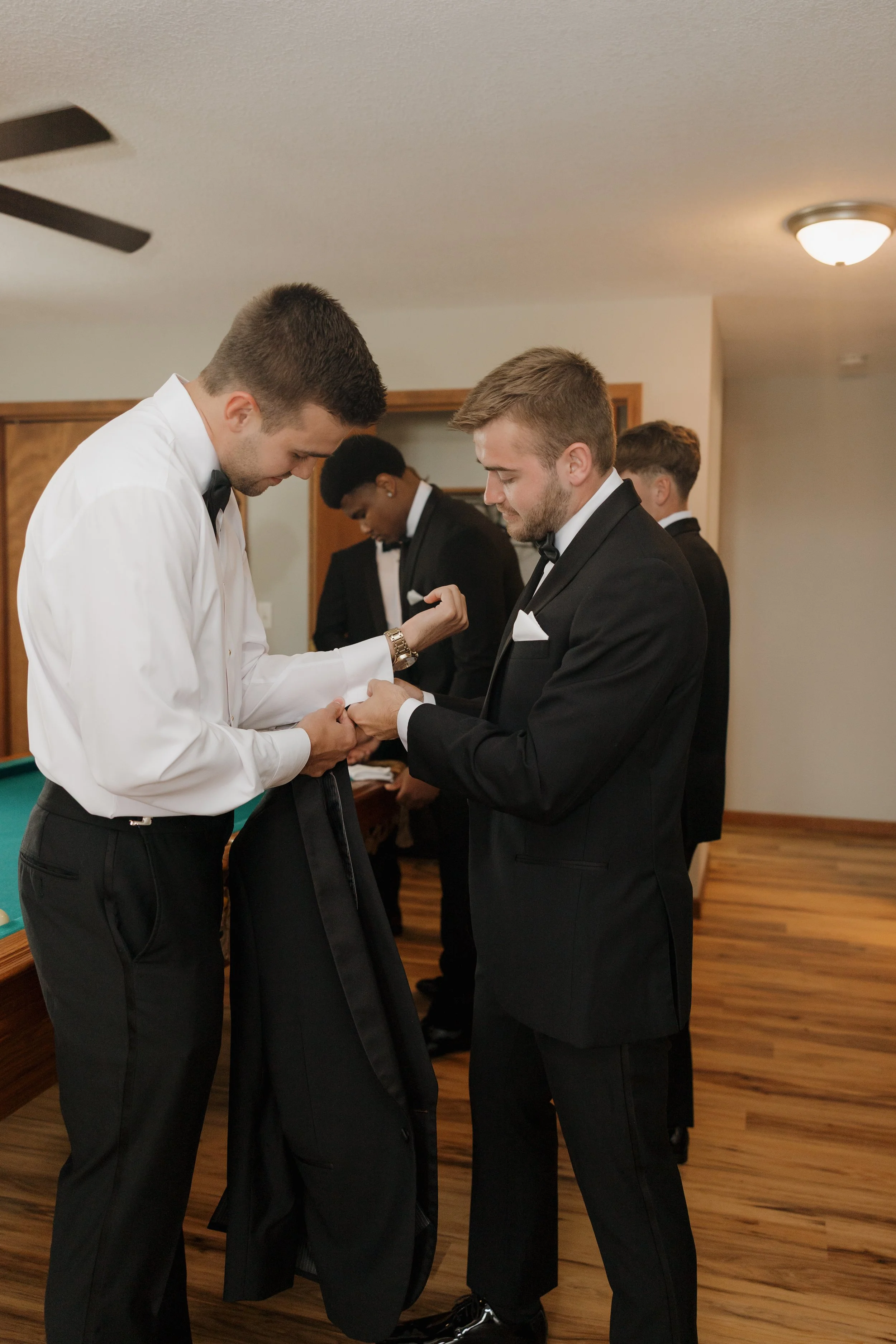 Four men in tuxedos preparing for a formal event in a room with wood flooring and a ceiling fan.