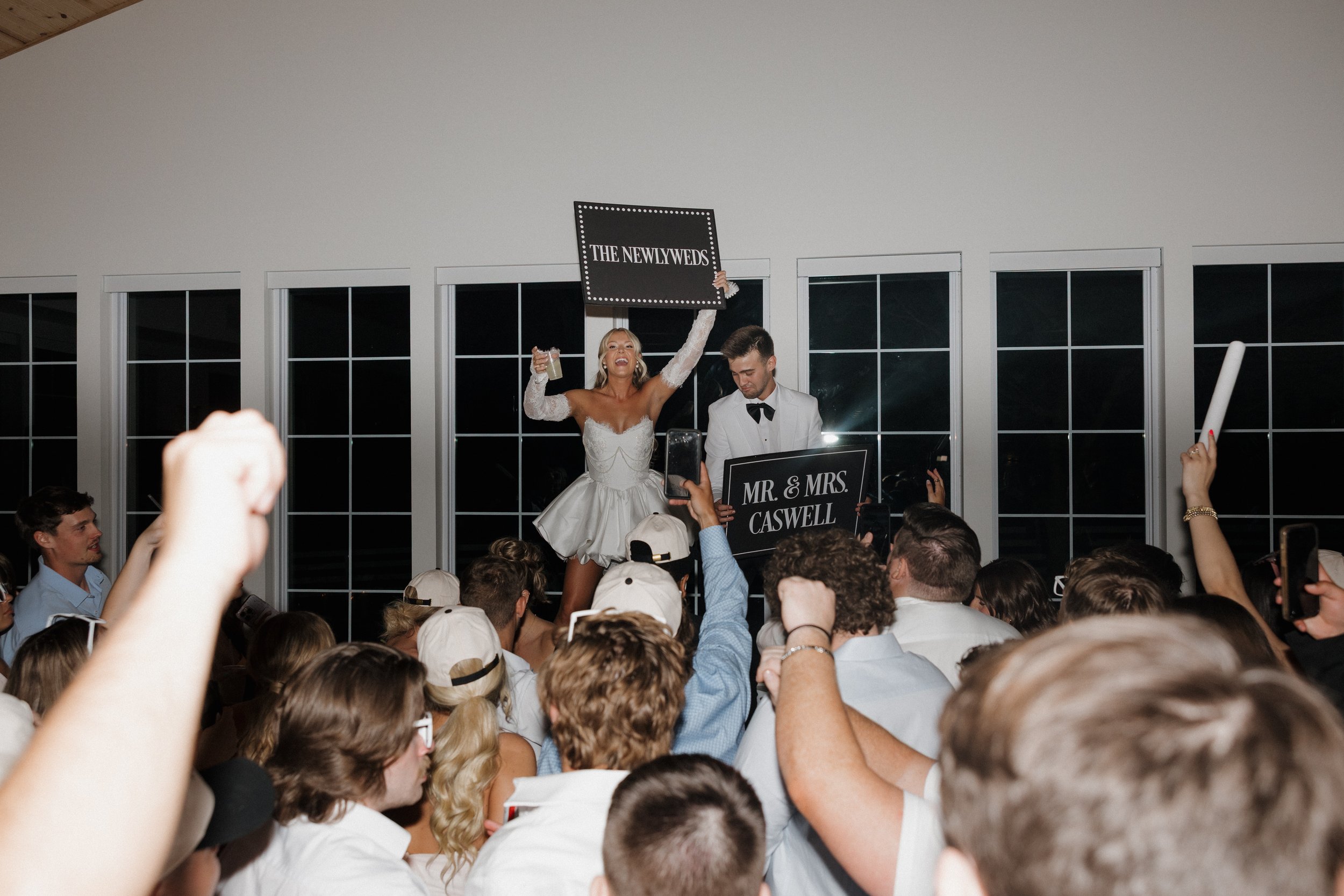 Wedding reception with bride in a white dress celebrating on stage while holding a sign, groom in tuxedo, surrounded by guests cheering, taking photos, and raising hands.