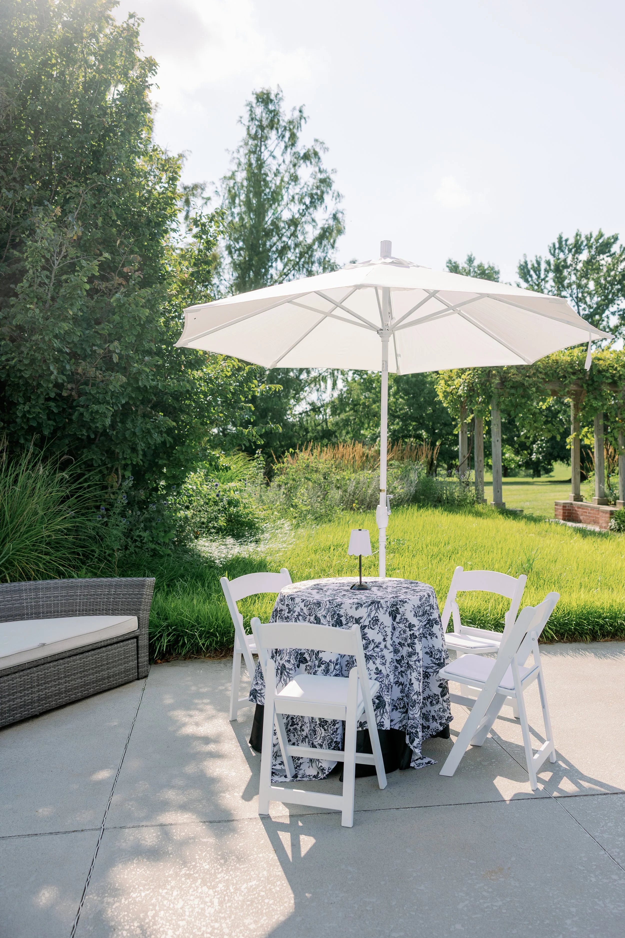 Outdoor patio table with a floral tablecloth, four white chairs, a white umbrella, and a small lamp on the table, set in a green garden with trees and a pergola in the background.