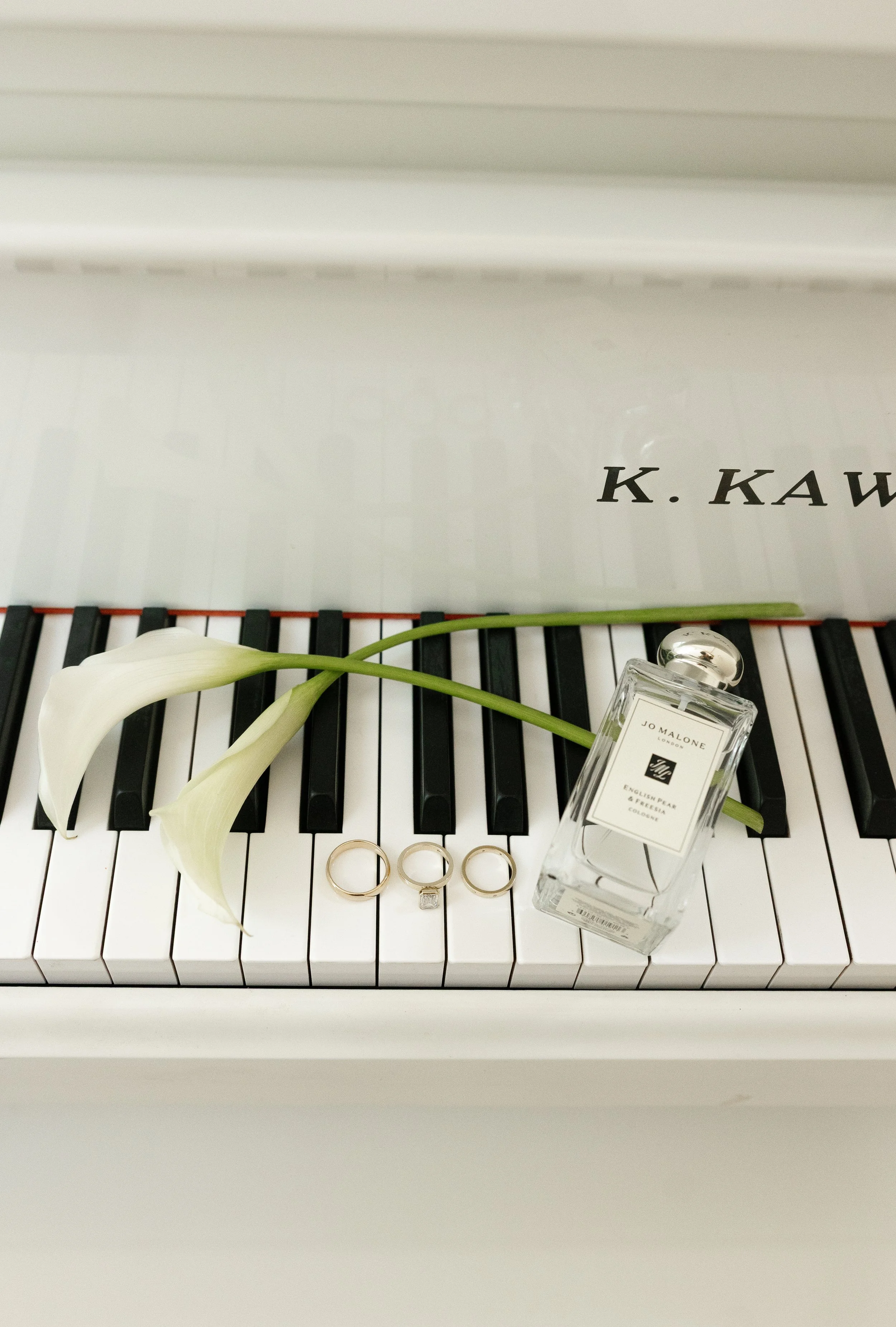 A white grand piano with the lid closed, featuring black and white keys. On the keys, there's a pair of calla lilies, three rings, a bottle of Jo Malone perfume, and a portion of text that reads 'K. KAW'.
