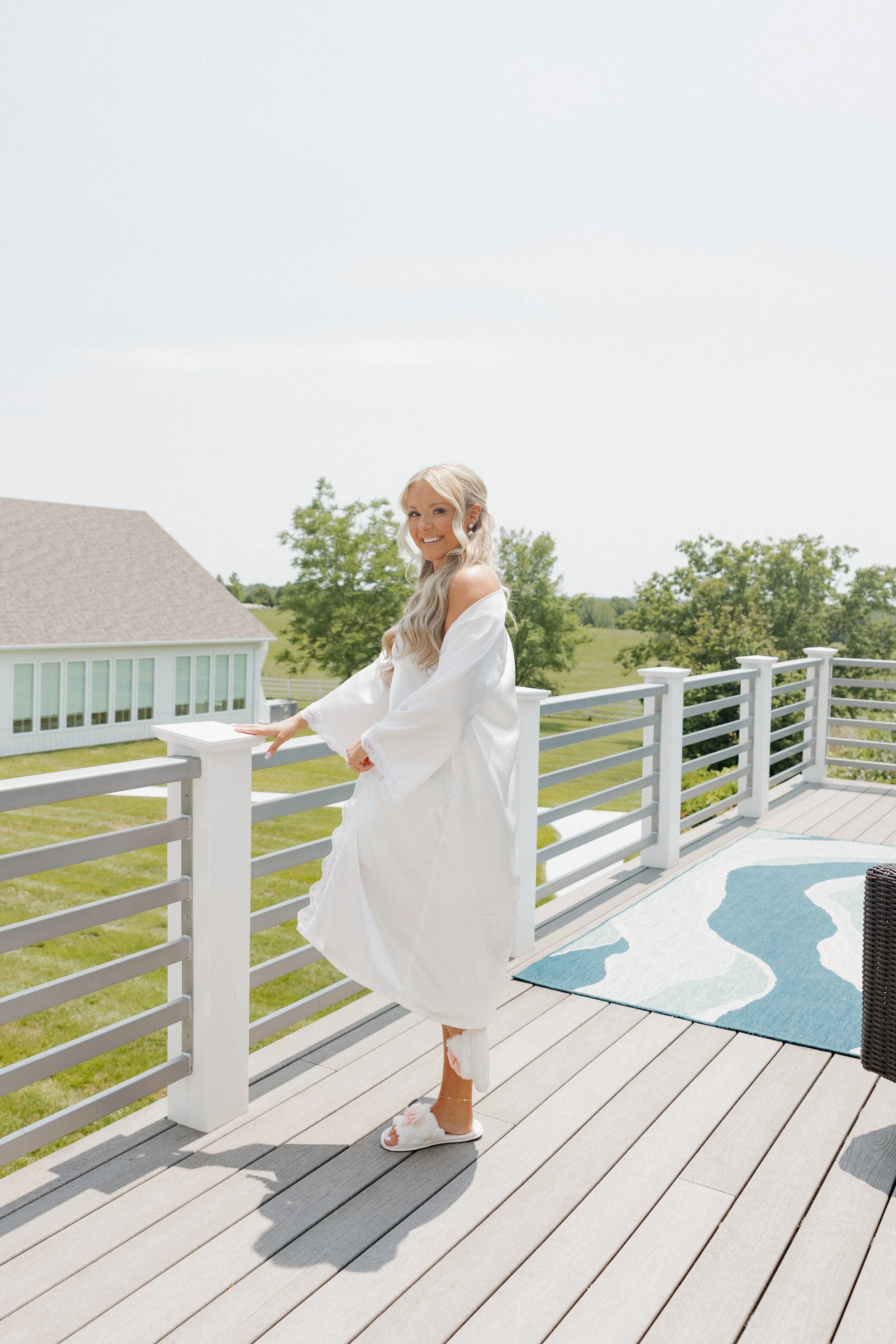 A smiling woman in a white robe standing on an outdoor deck with a railing, overlooking a grassy field and trees under a clear sky.