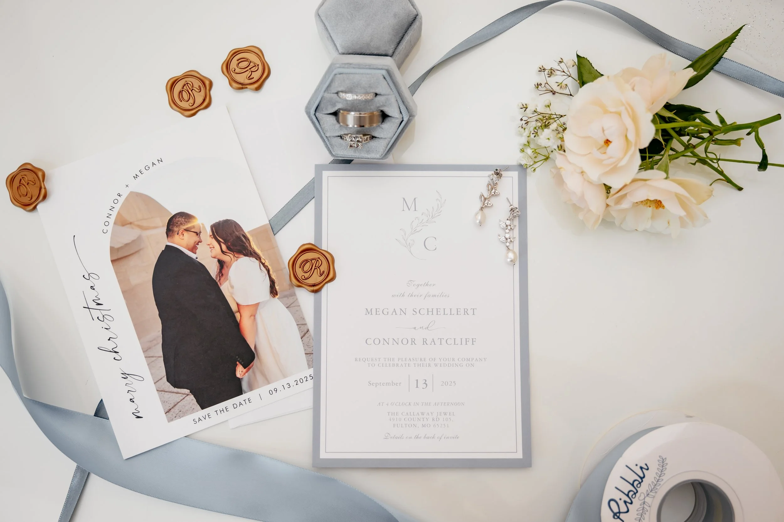 Wedding invitation, a 'Save the Date' card featuring a photo of a couple, wedding rings in a gray velvet box, pearl earrings, cream flowers, and sealing wax stamps with initials, all arranged on a white surface.