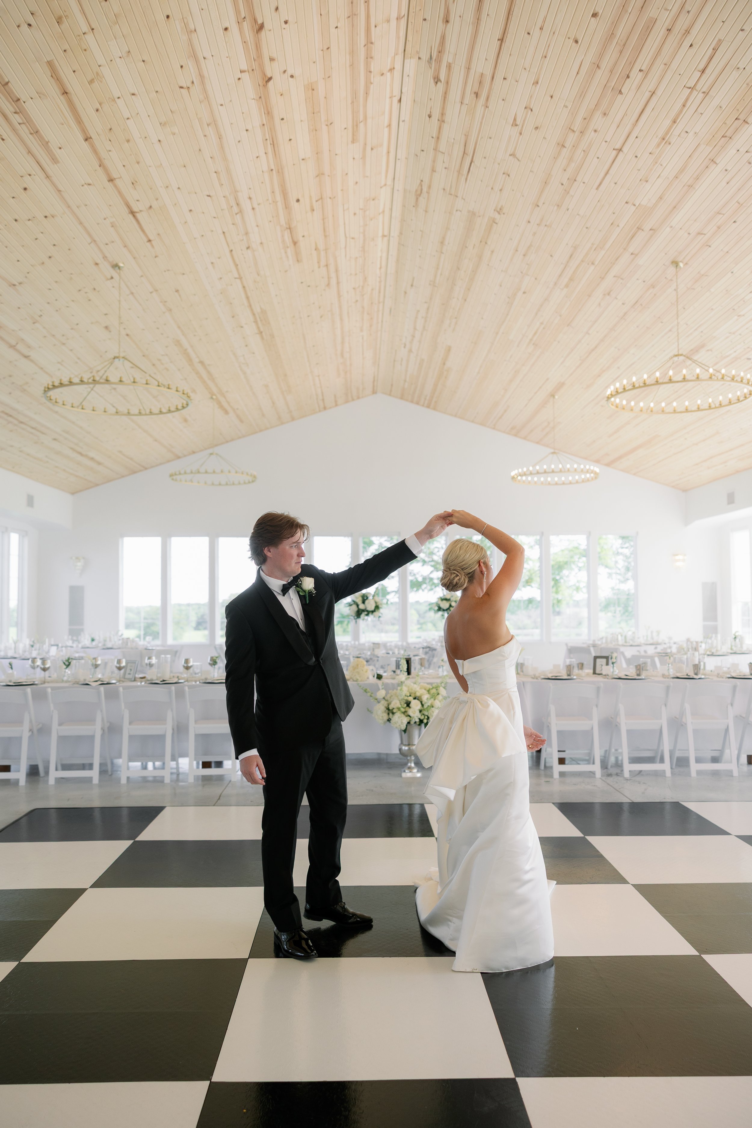 A bride and groom dancing in a wedding reception hall with a checkered black and white floor, surrounded by tables with white chairs and floral centerpieces, under a wooden vaulted ceiling with circular chandeliers.