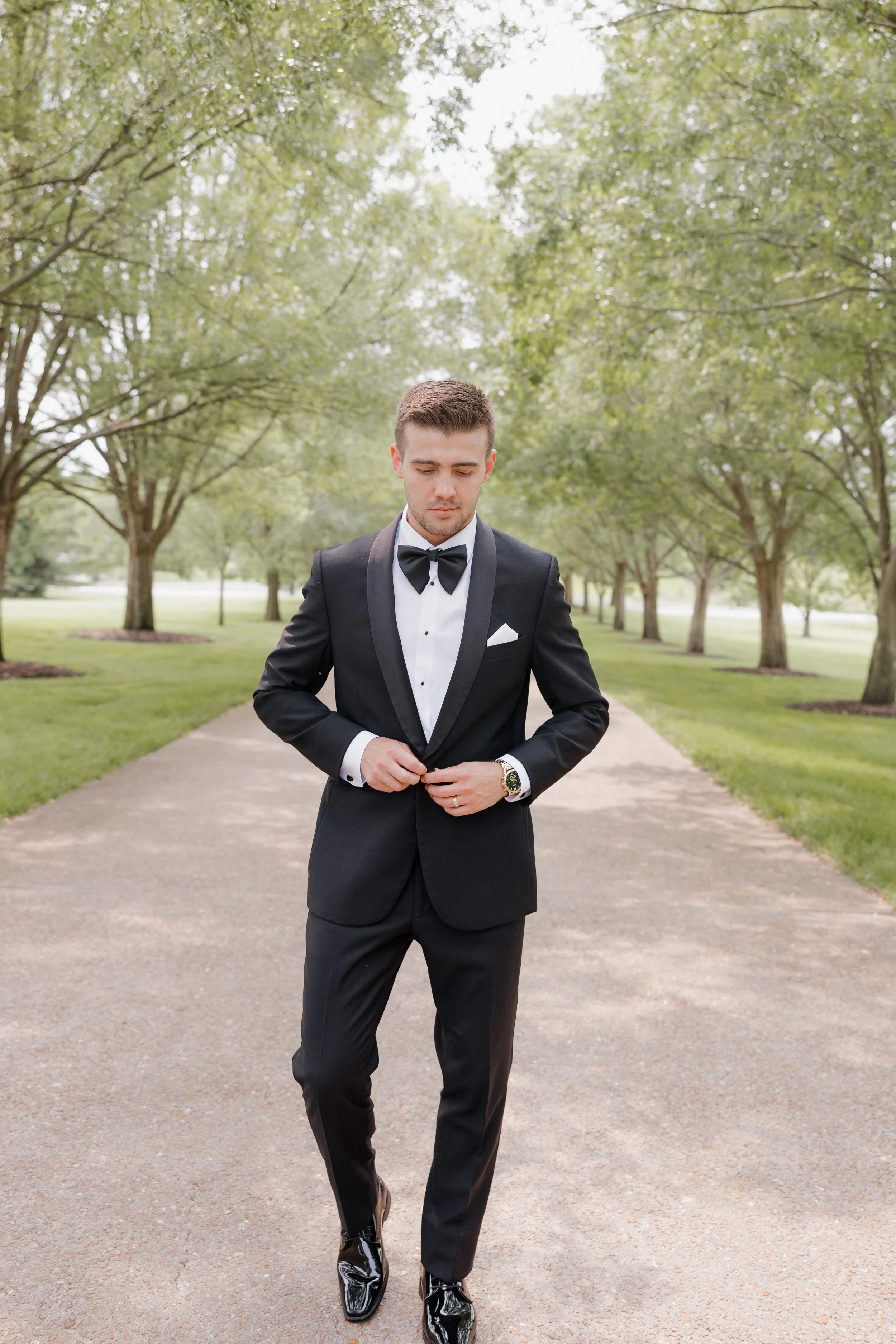 A young man in a black tuxedo with a bow tie, standing on a walkway in a green park, adjusting his jacket.