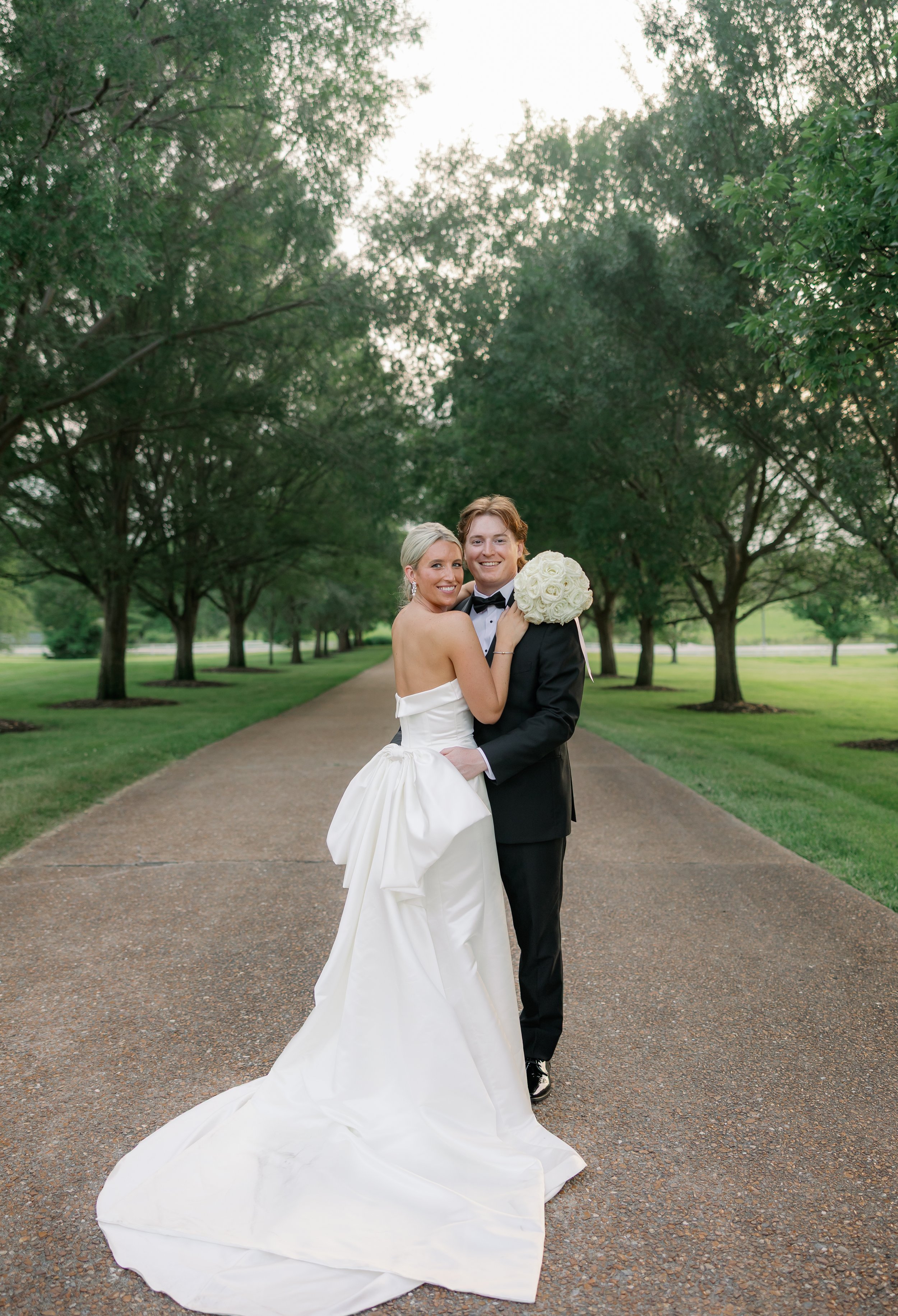 A newlywed couple hugging on a tree-lined pathway, bride in white wedding gown holding bouquet, groom in black tuxedo, during daytime.