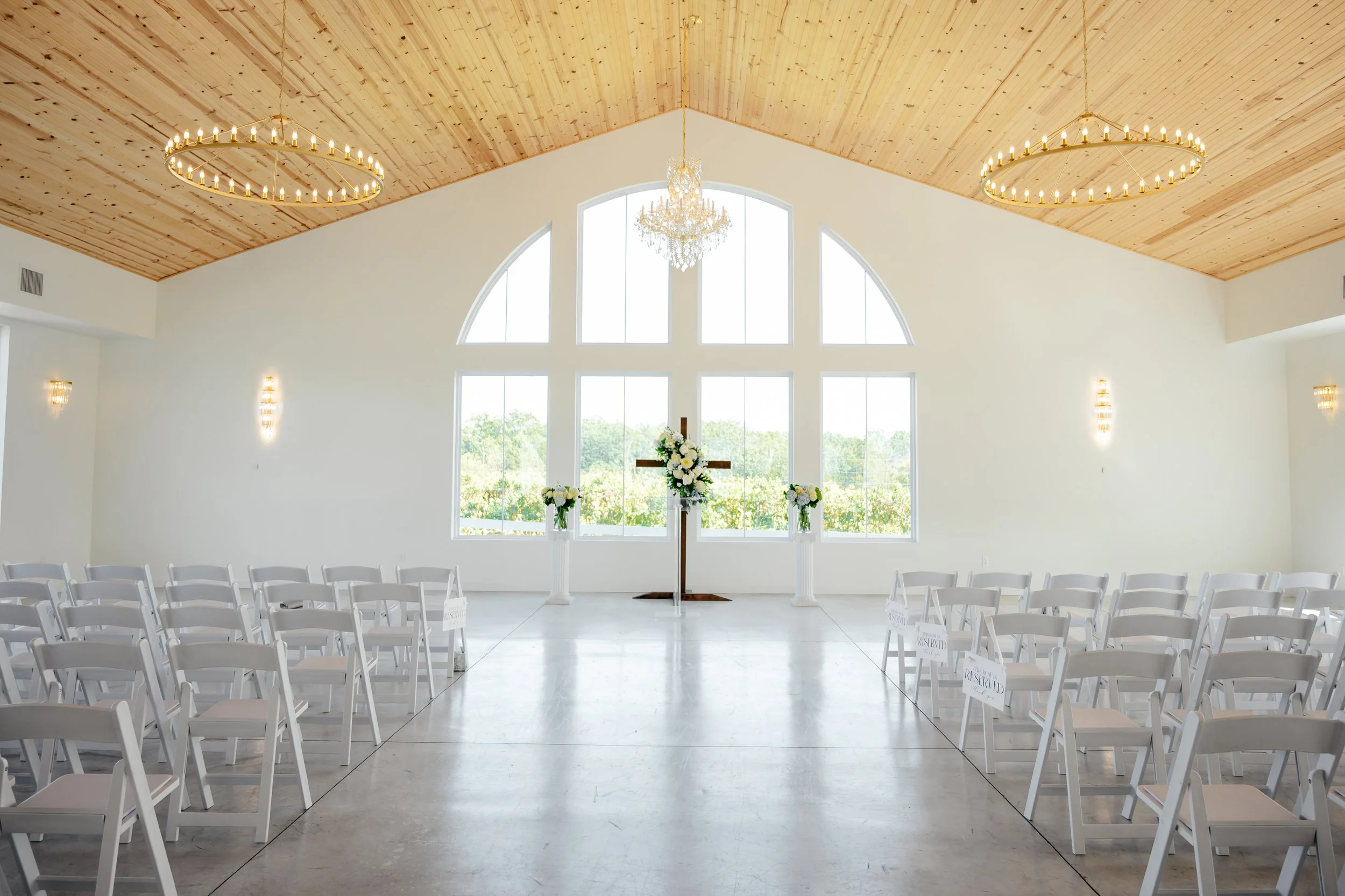 Empty wedding ceremony room with white chairs, floral arrangements, large arched window, and a wooden cross at the altar.