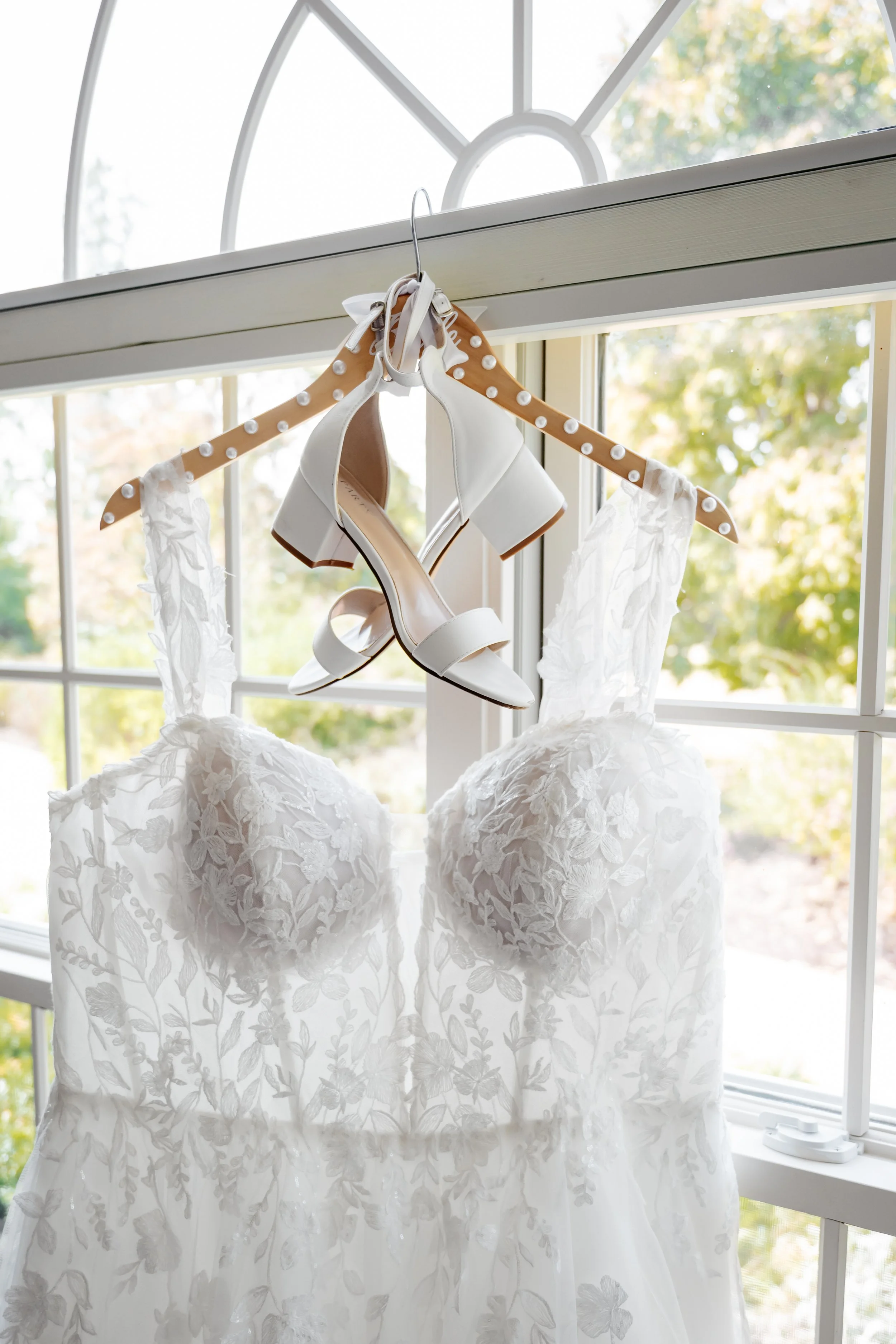 A white wedding dress with floral lace details hung on a hanger in front of a window, with a pair of white high-heeled shoes also hanging on the hanger.